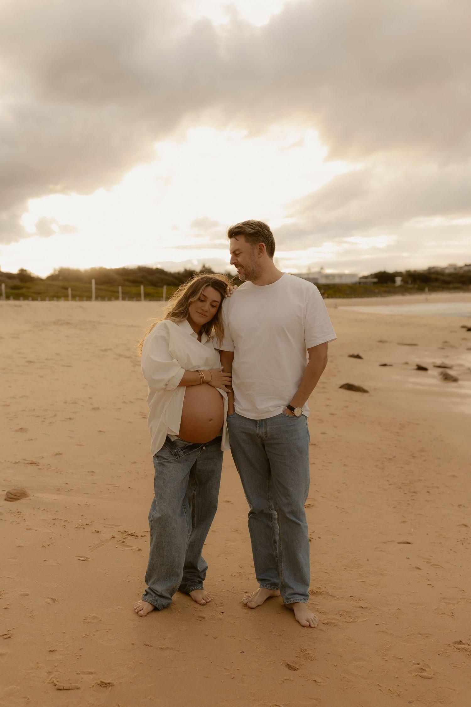 Couple on beach at sunset, pregnant woman in white shirt and jeans with partner in white tee, maternity photo shoot.