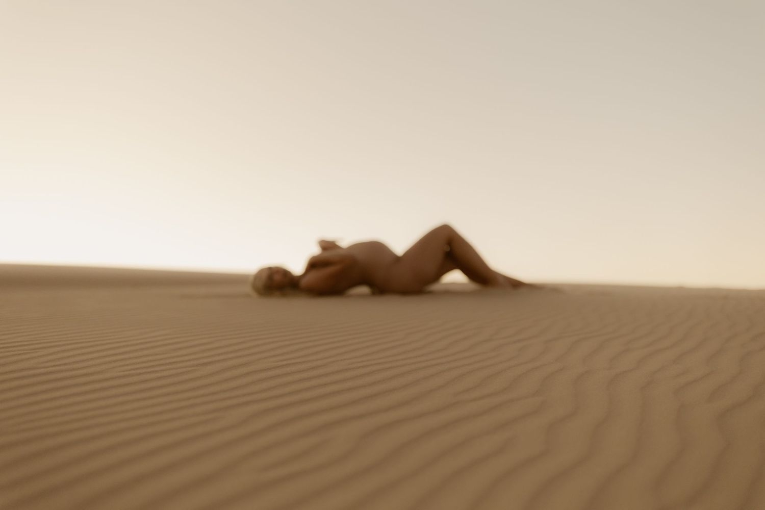 A nude figure lies gracefully on rippled golden sand dunes in a vast desert landscape under a hazy sky.