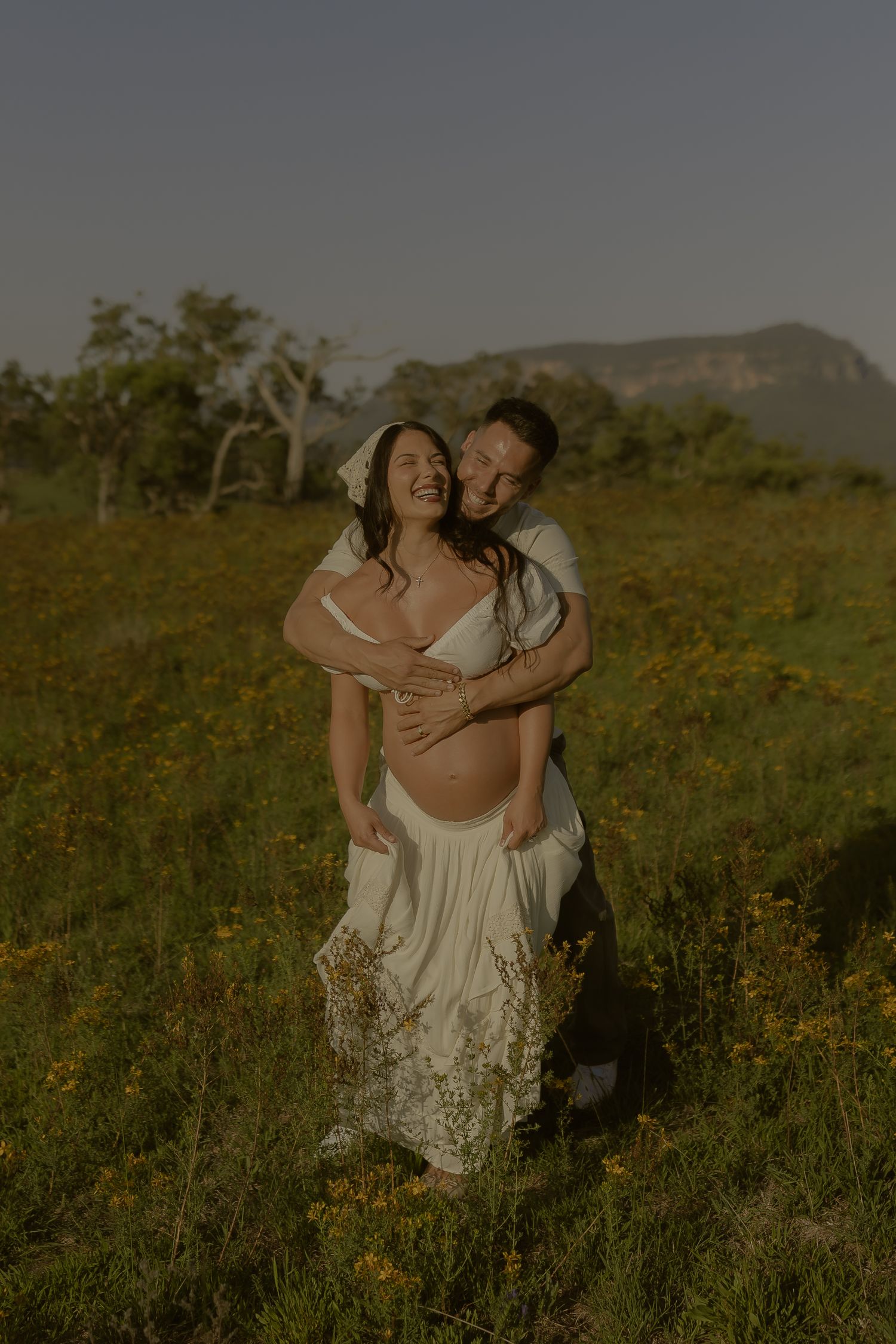 A couple embraces in a sunlit wildflower field wearing white outfits during a romantic photoshoot.