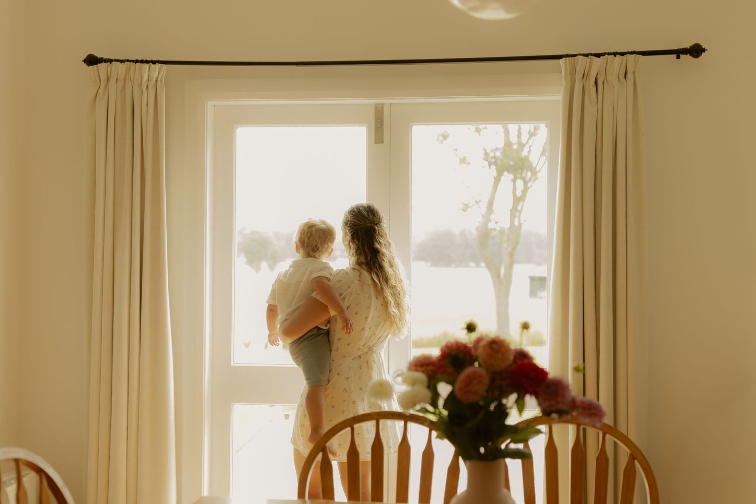 A bright interior room with sunlit windows, wooden dining furniture and flowing curtains.