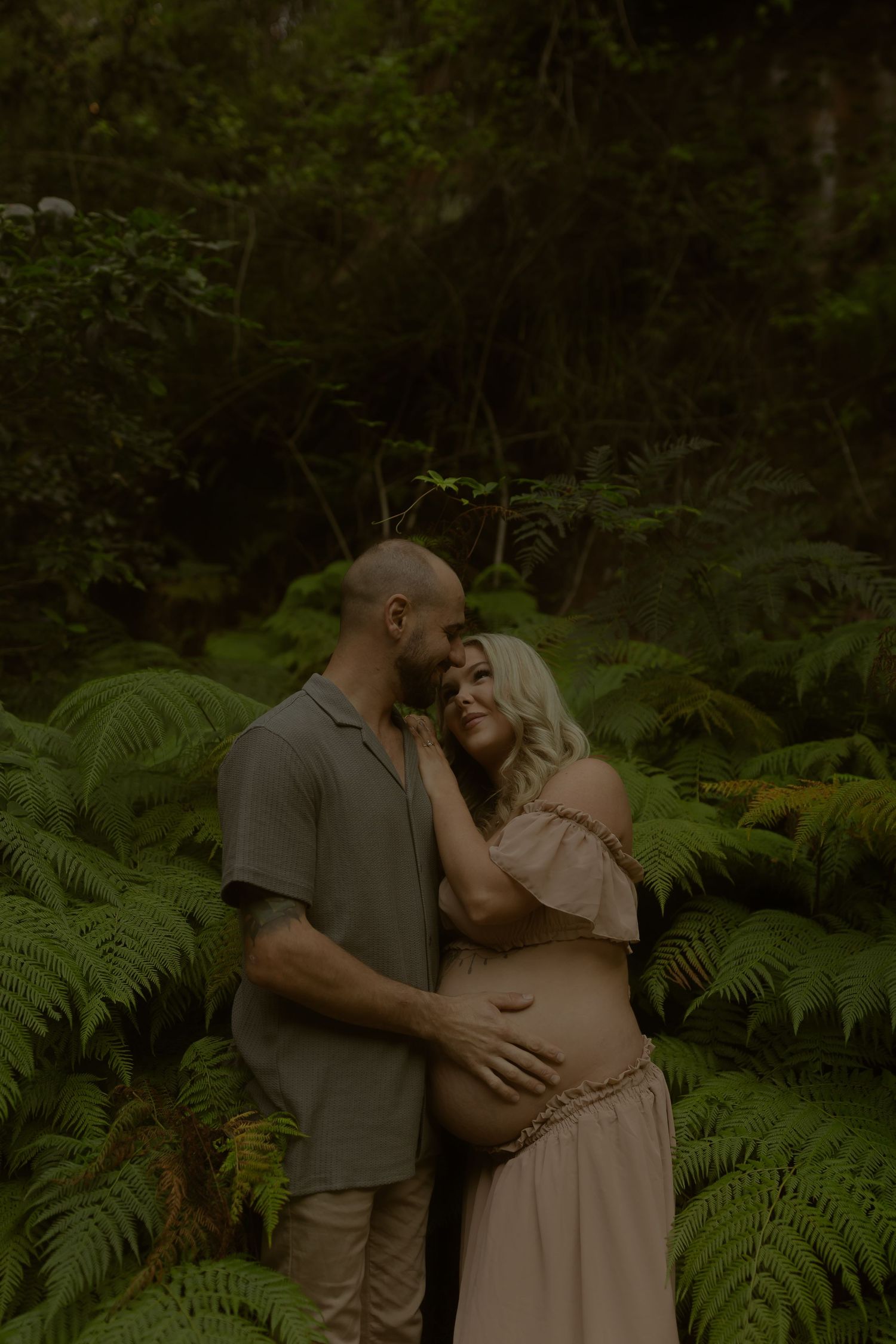 Couple embracing in lush fern forest, pregnant woman in beige dress with partner in olive shirt.