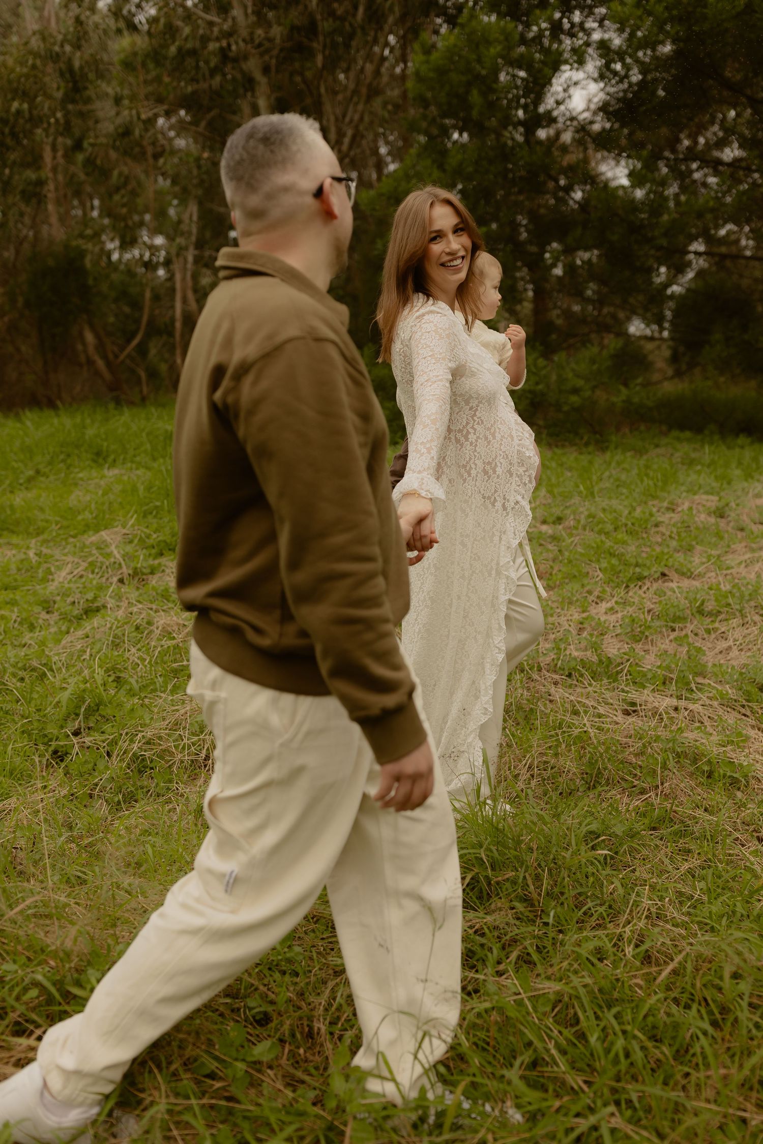 Couple walking hand in hand through green field, bride in lace gown looks back smiling, groom in olive jacket and white pants.