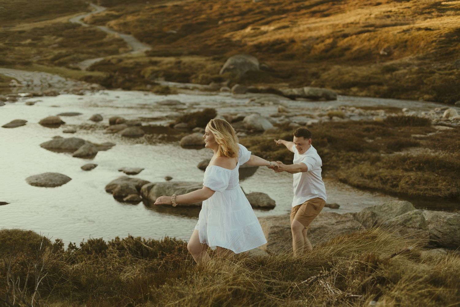 Couple in white clothing dances together at sunset on rocky coastal terrain.