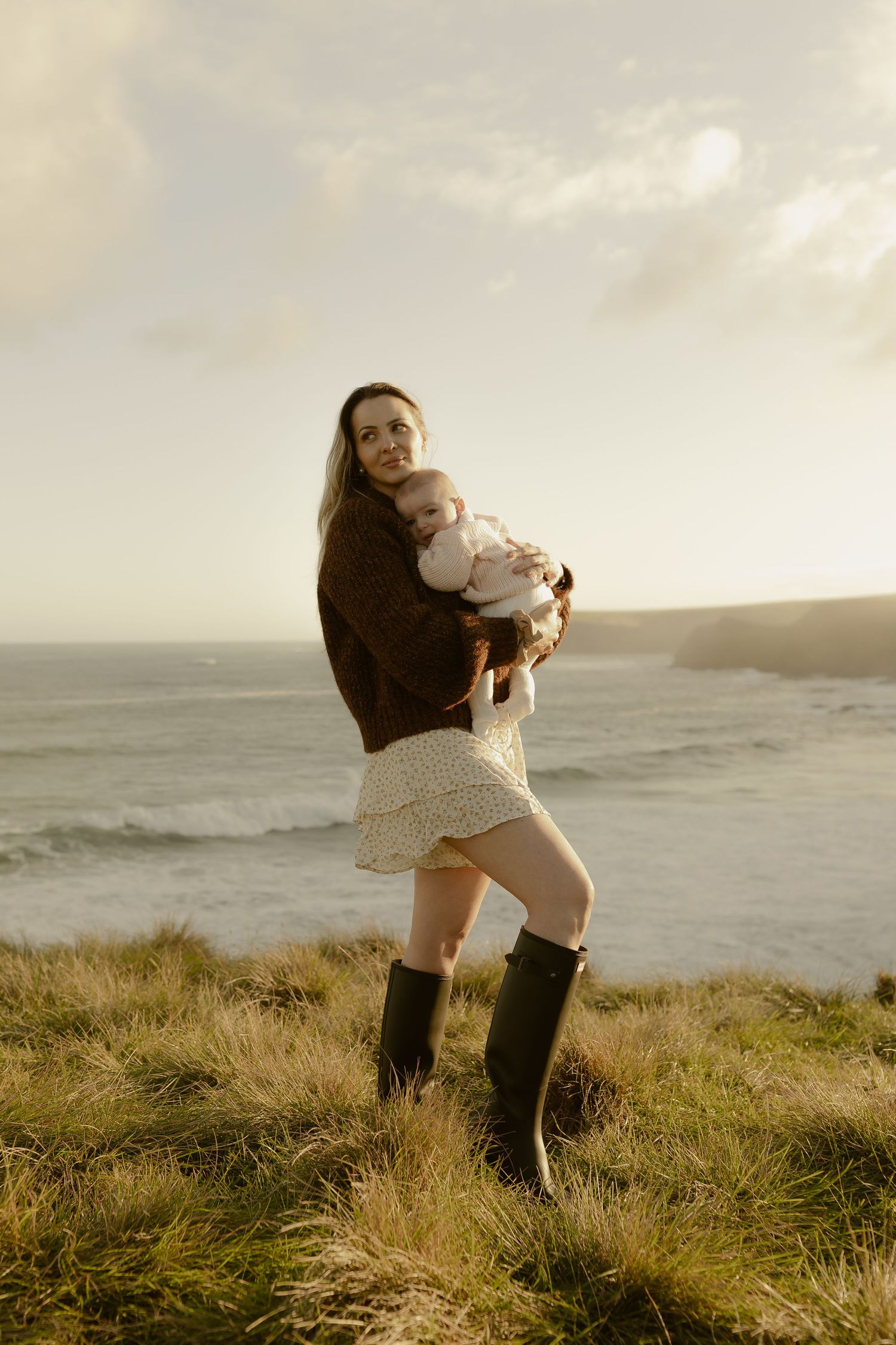 Woman in brown jacket and rubber boots holds baby on grassy coastal cliff with ocean backdrop.