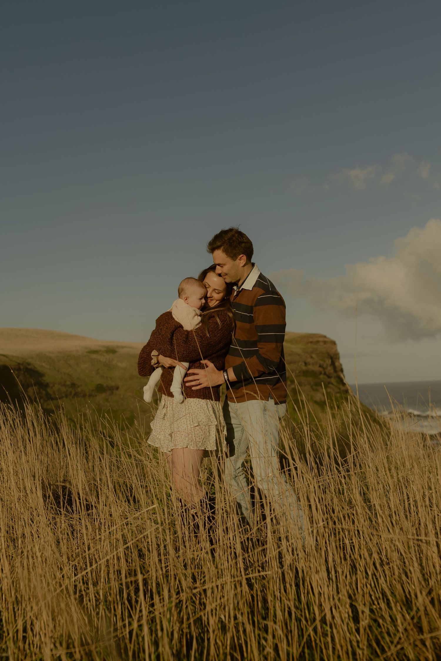 Couple embracing in golden tall grass with coastal cliffs and blue sky backdrop.