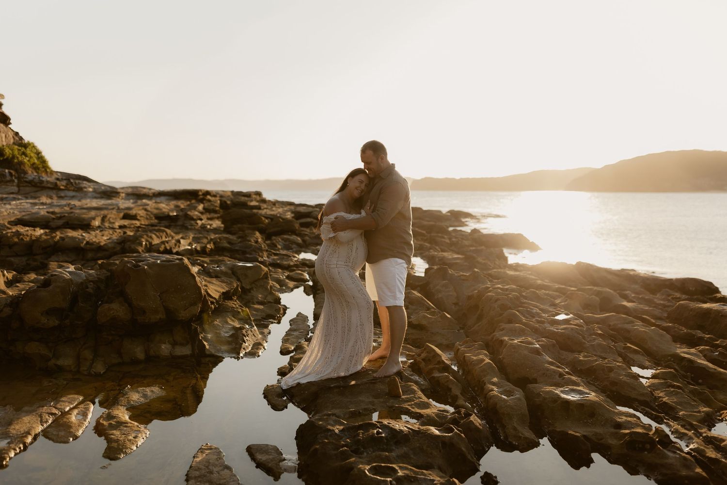 A couple poses on rocky coastal cliffs at sunset with ocean waves crashing below.
