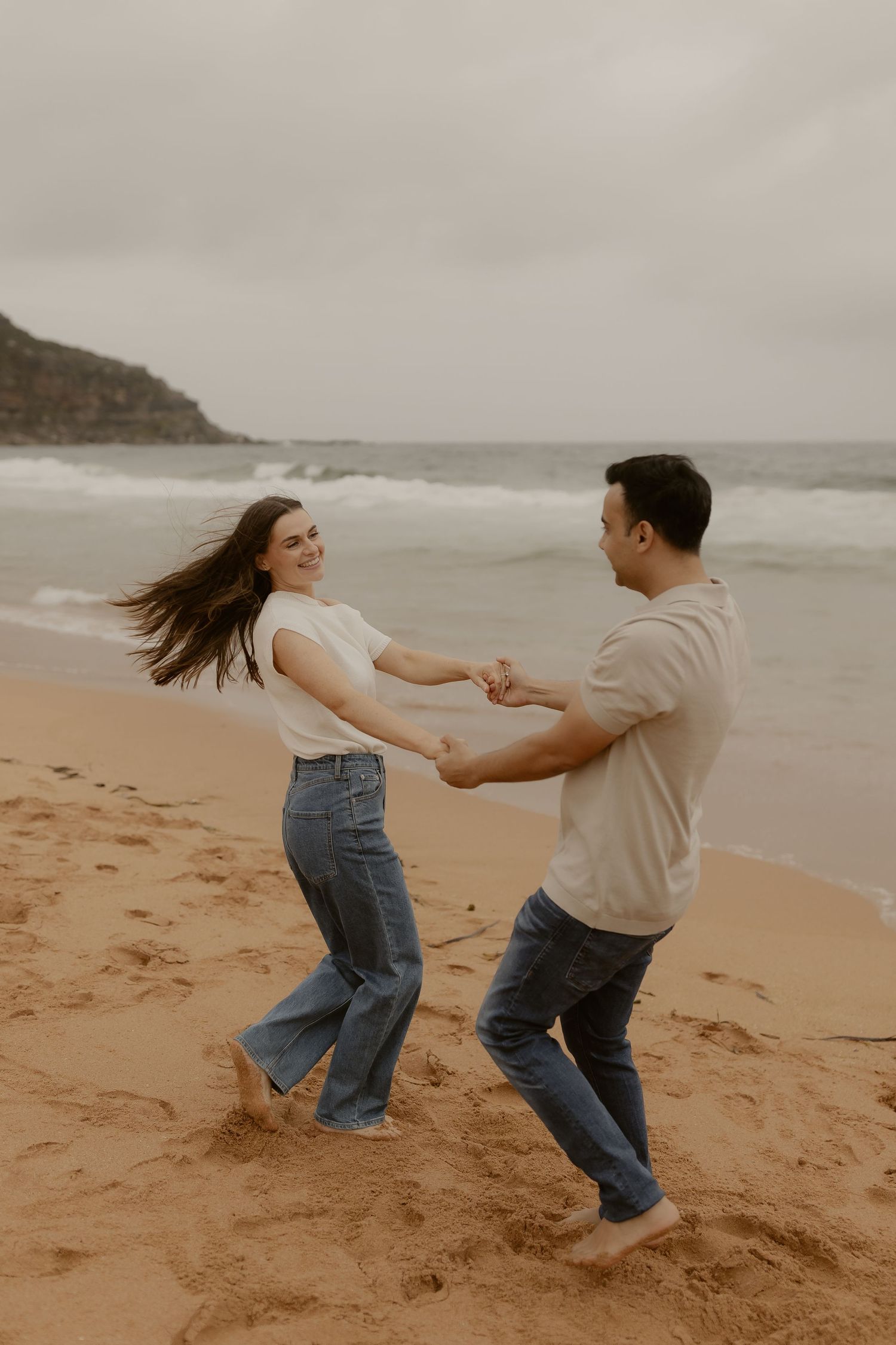 A playful couple spins and dances together on a sandy beach by the ocean.