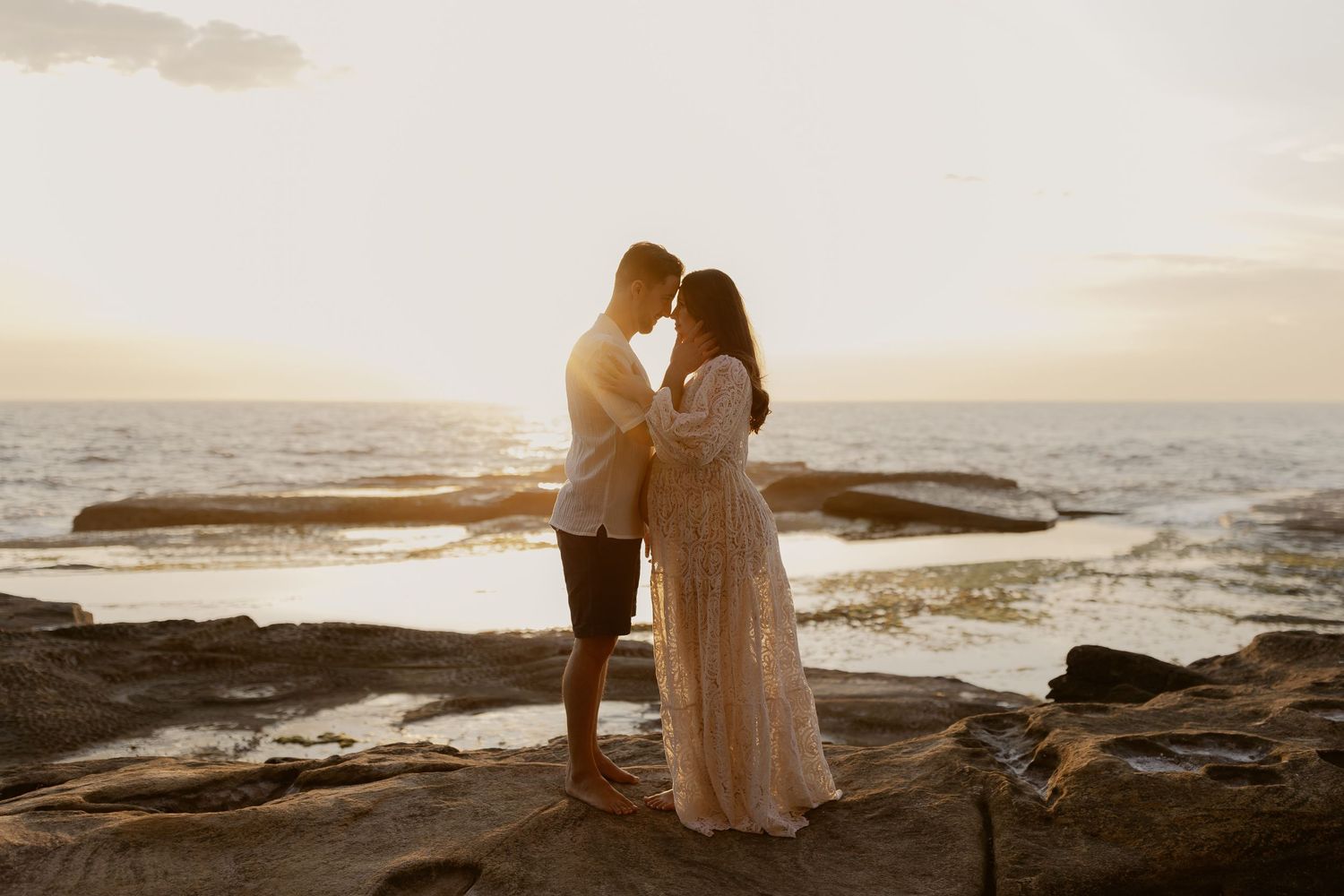 Couple in romantic embrace on rocky coastline at sunset with ocean waves crashing in background.