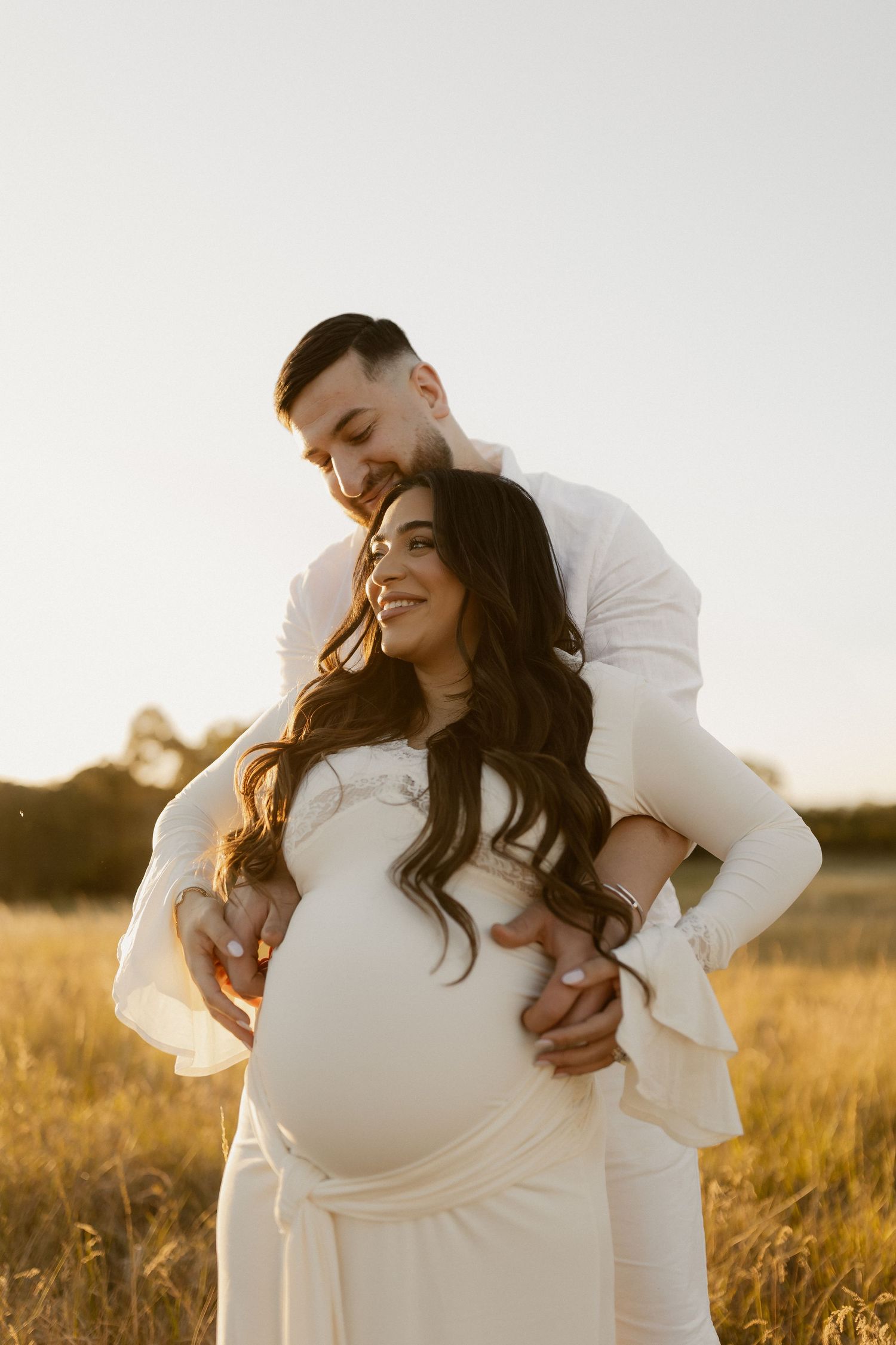 An intimate maternity photoshoot in a sun-drenched field captures a tender moment between expectant parents.