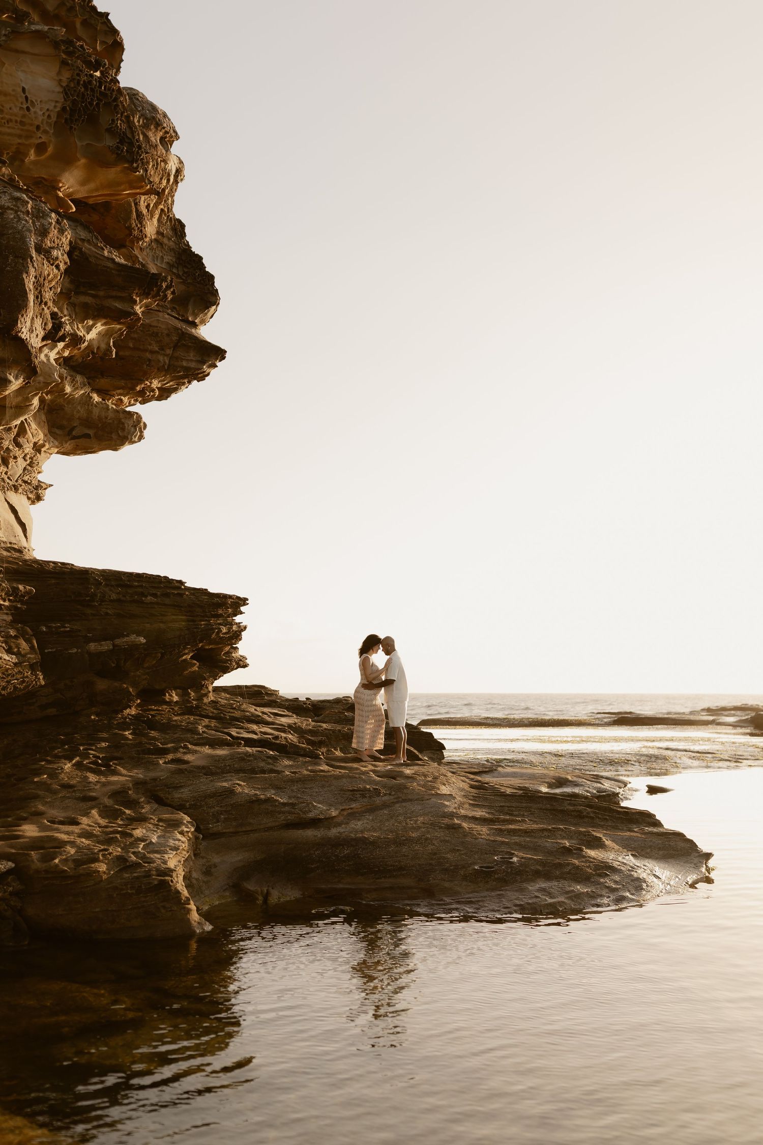 Silhouetted couple embracing on rocky coastal cliffs at sunset with ocean waves below.