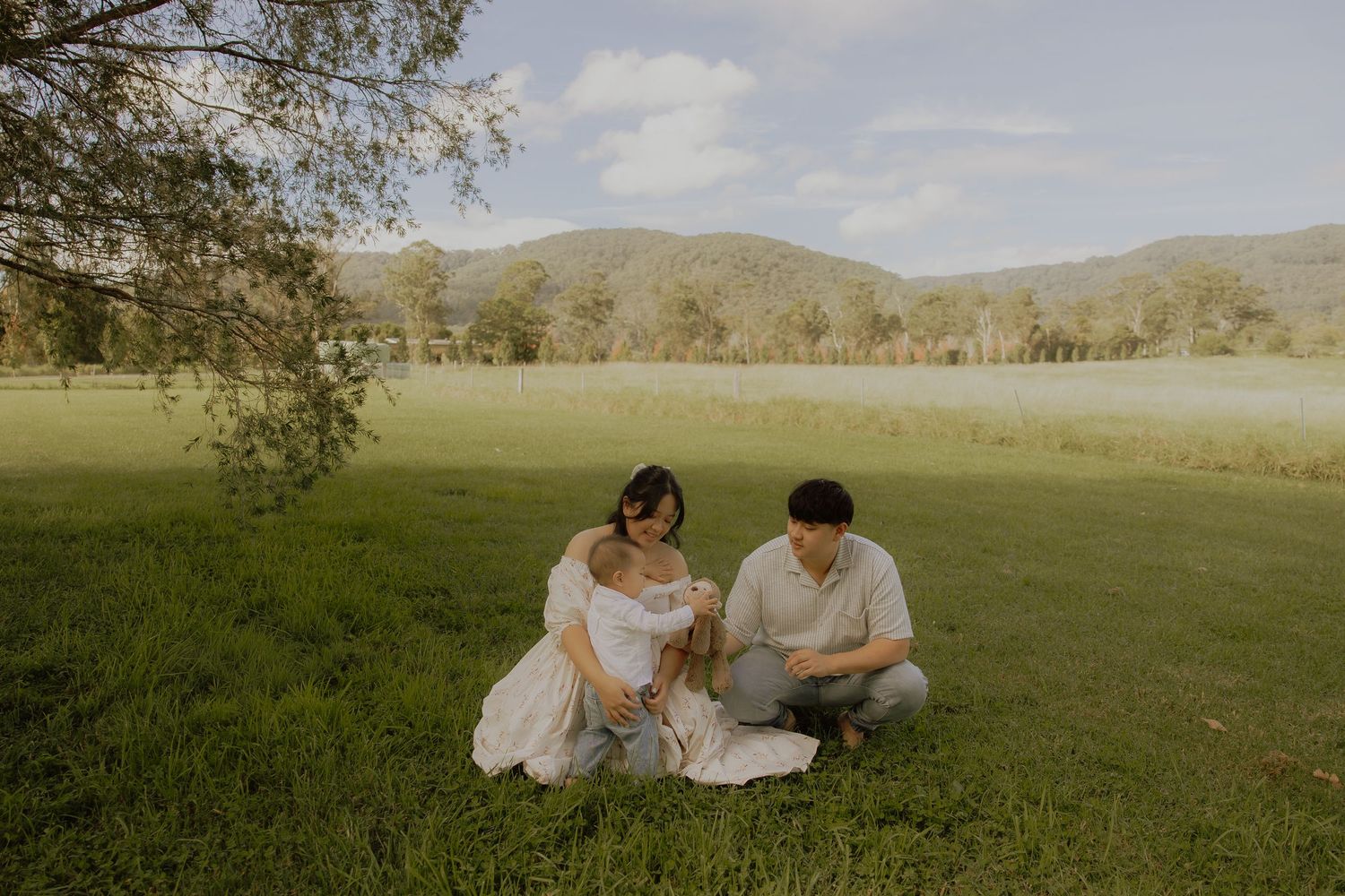 Couple sitting on lush green grass with mountains in background during outdoor maternity photoshoot.