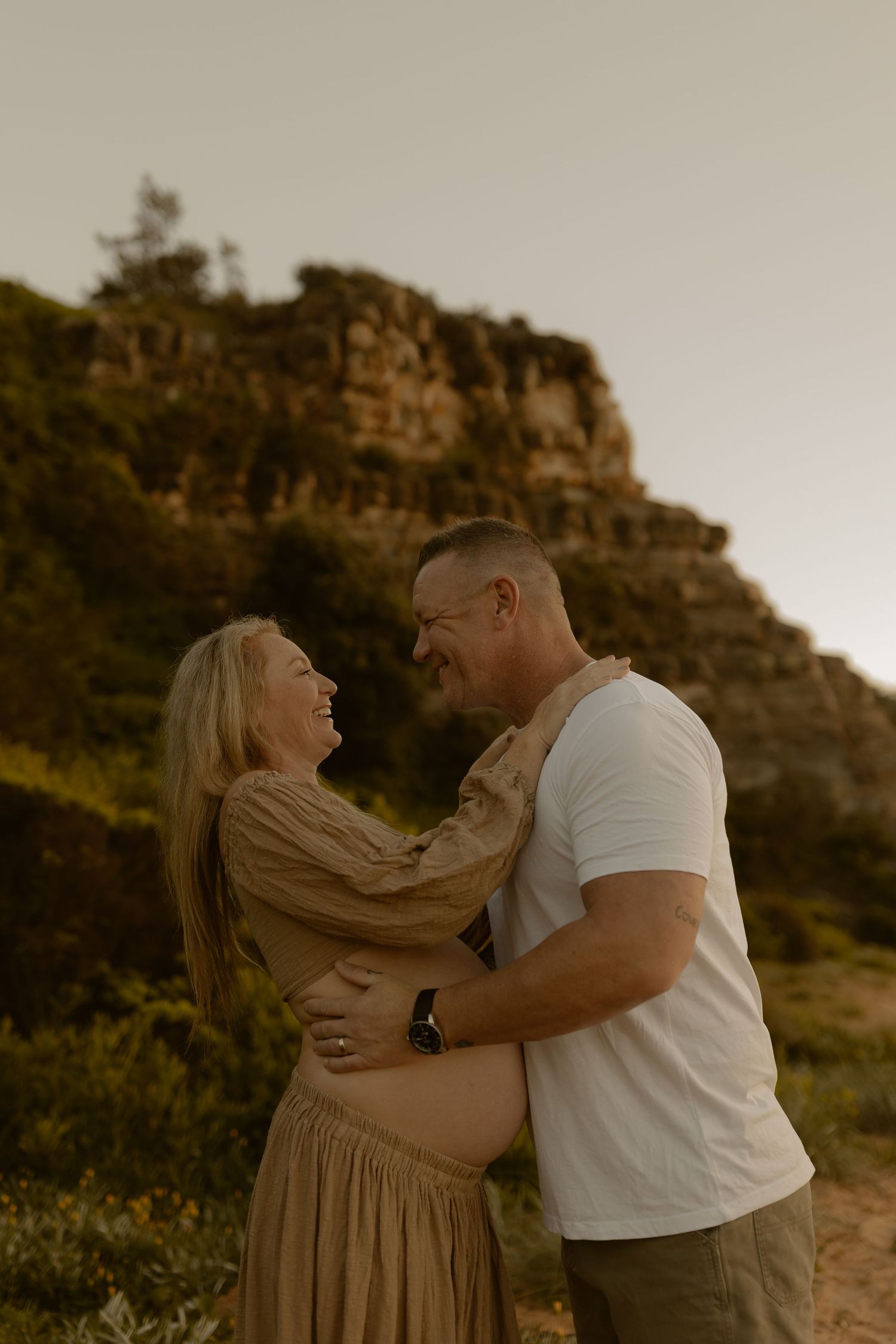 A couple embraces and laughs together outdoors with rocky cliffs and golden light in the background.