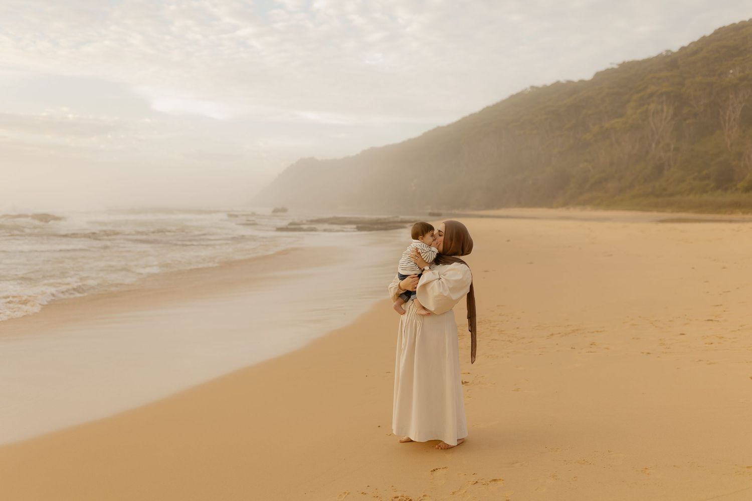 A mother holds her child on a sandy beach at sunset, with ocean waves and a green hillside in the background.