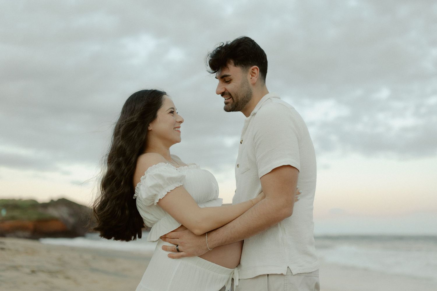 A couple embraces on a cloudy beach at sunset, wearing white and beige outfits.