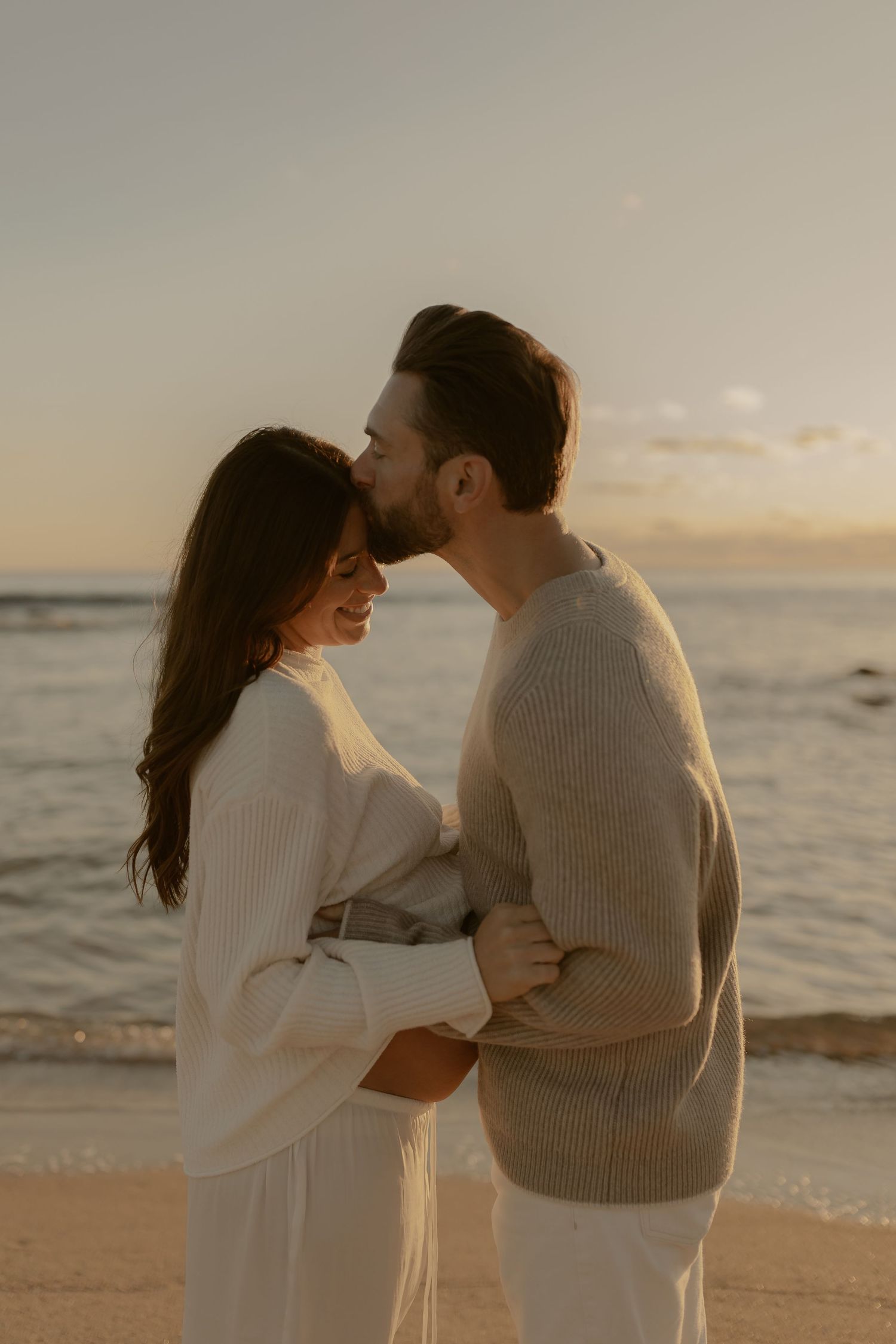 A couple in cozy sweaters embraces tenderly on a beach at sunset while waves crash behind them.