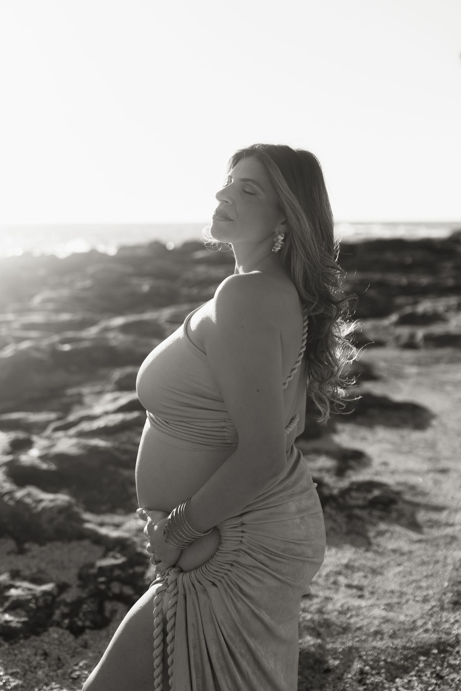 Black and white maternity photo of pregnant woman in draped dress posing on rocky beach at sunset.