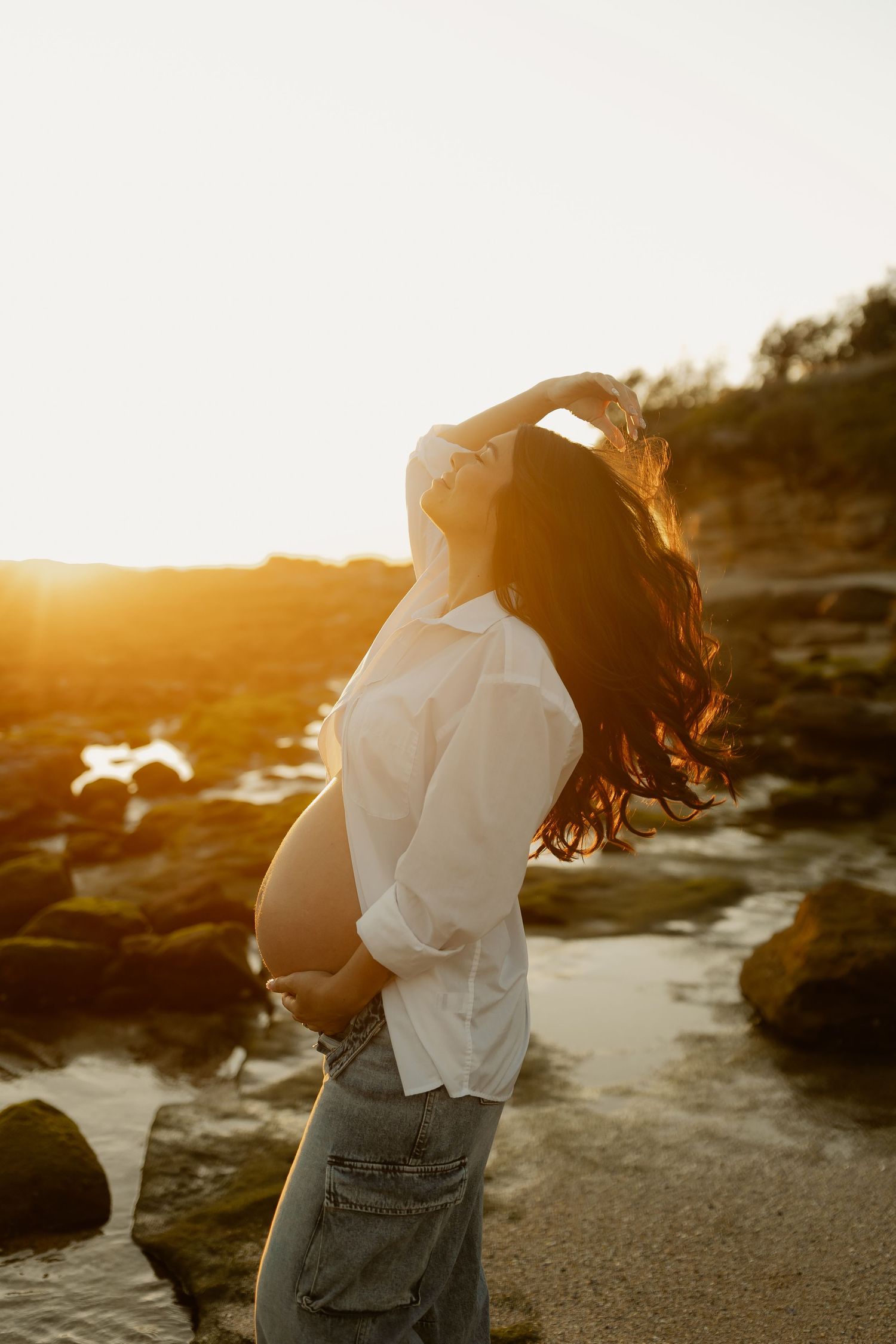 Pregnant woman tossing hair back on rocky beach, holding bump during glowing golden sunset.