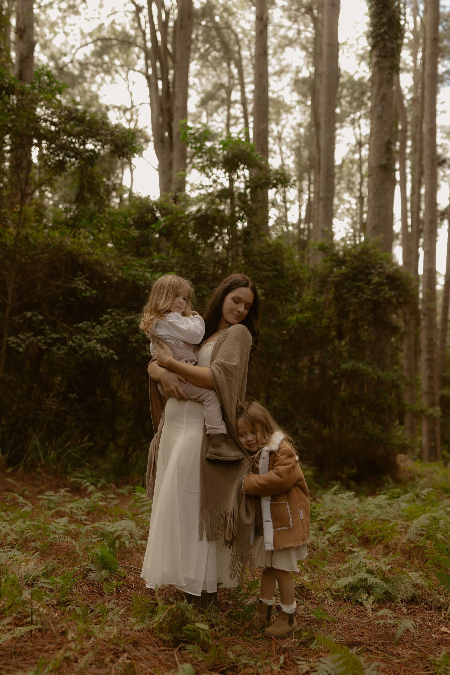 A mother in a white dress holds a toddler while another child hugs her leg in a lush forest setting.