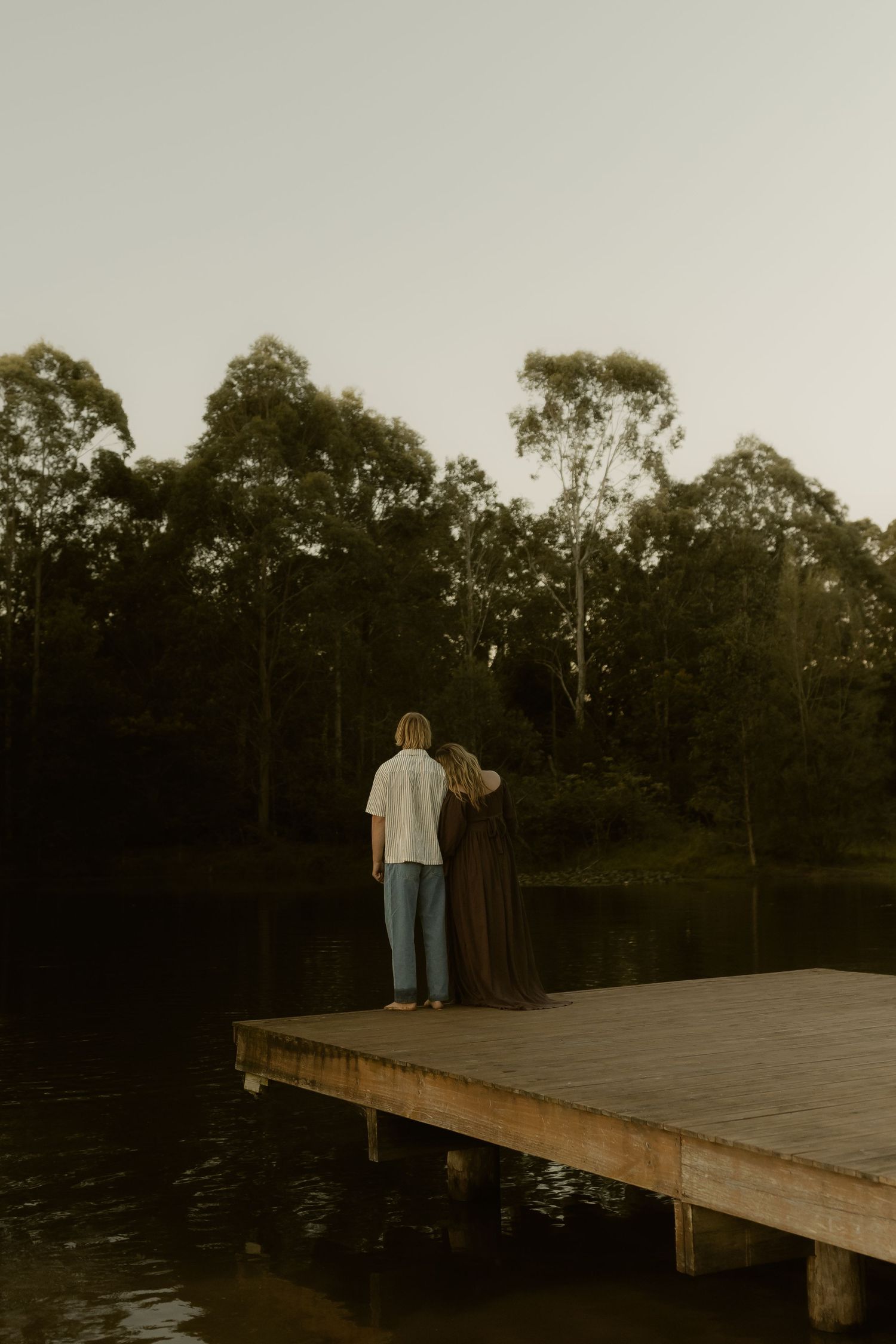 Couple standing together on a wooden dock overlooking a tranquil lake surrounded by tall eucalyptus trees at dusk.