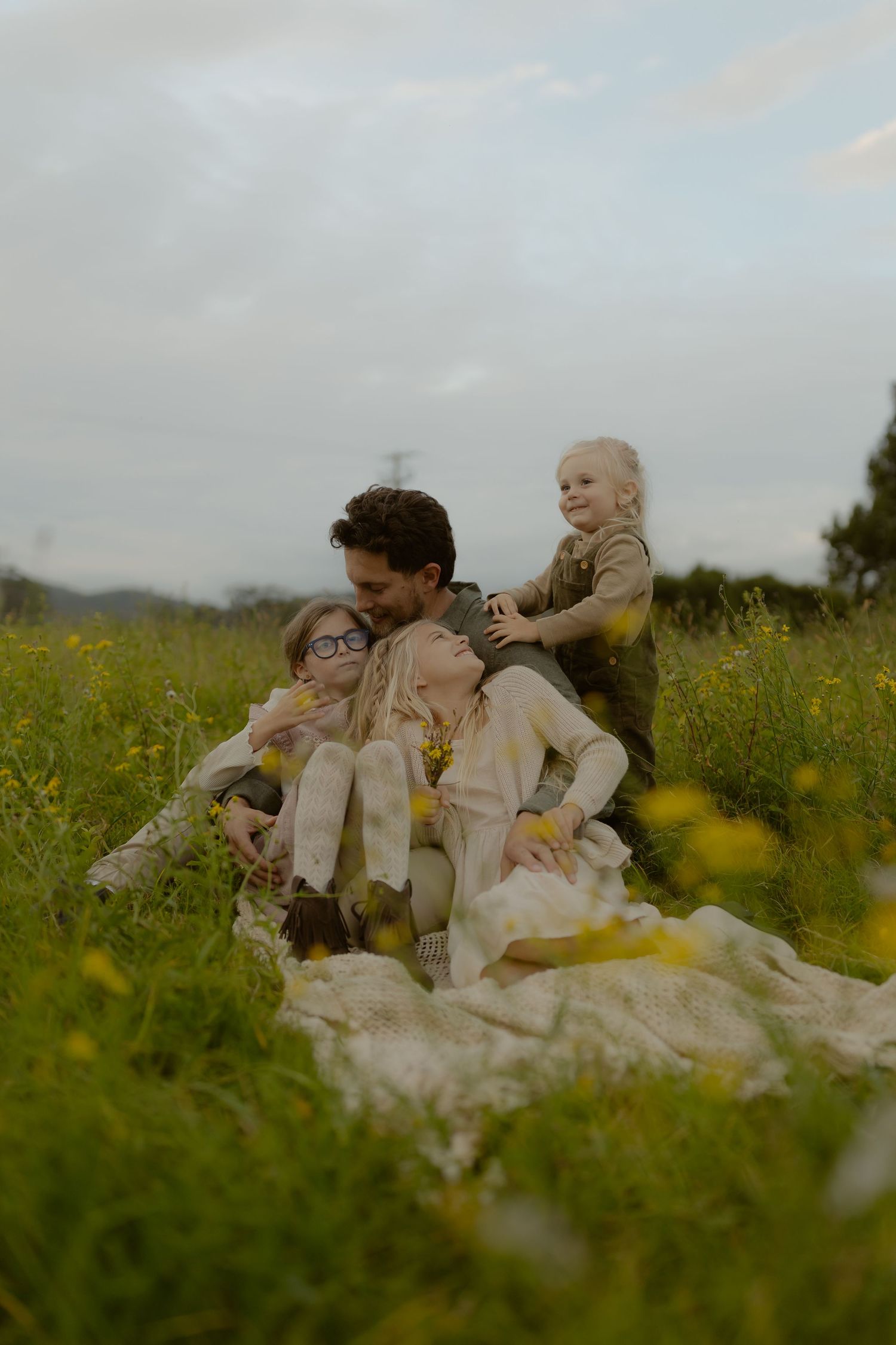 A family of four sits together on a blanket in a field of yellow wildflowers under a cloudy sky.
