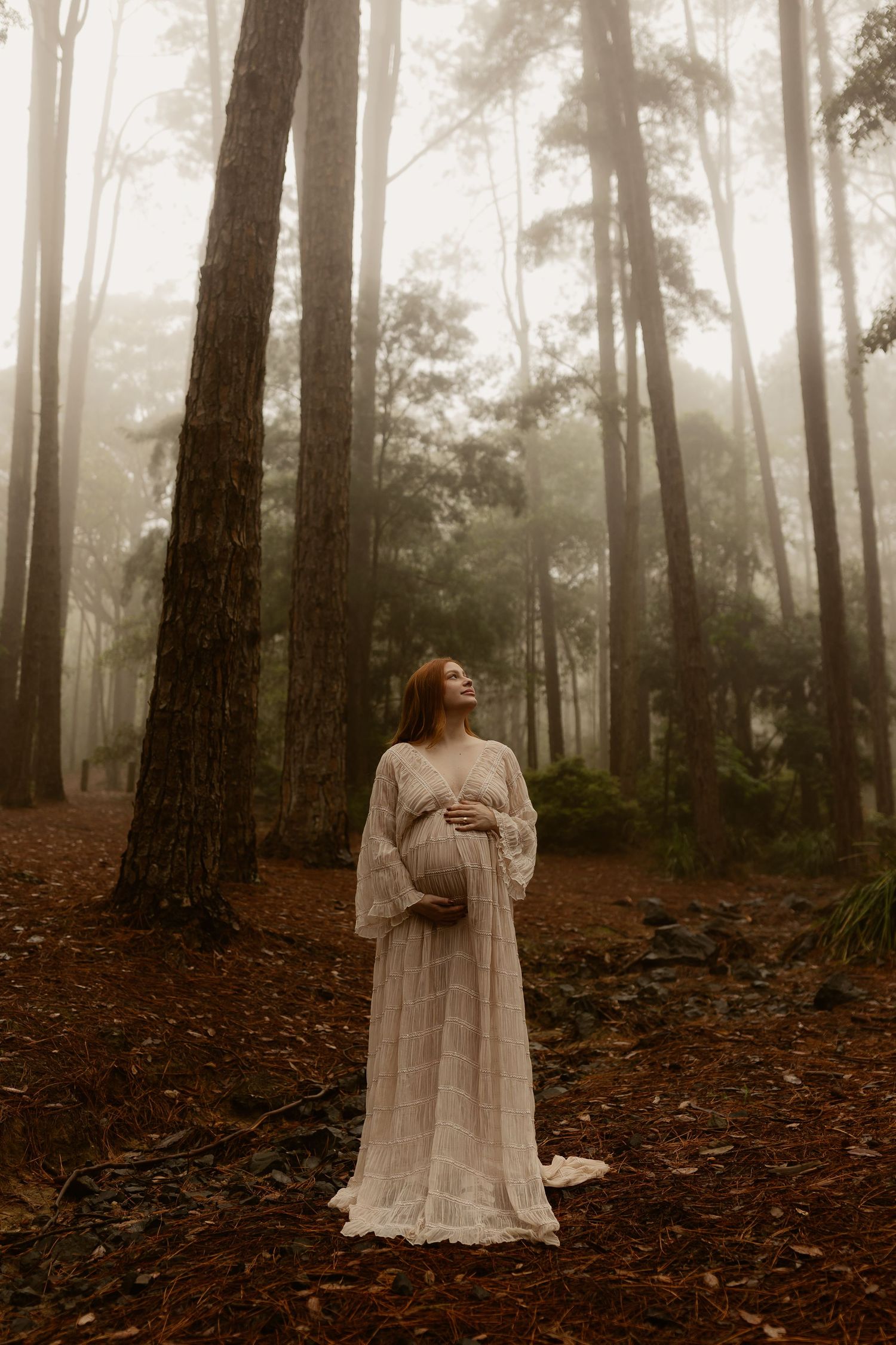 Pregnant woman in white lace gown standing among tall trees in foggy forest.