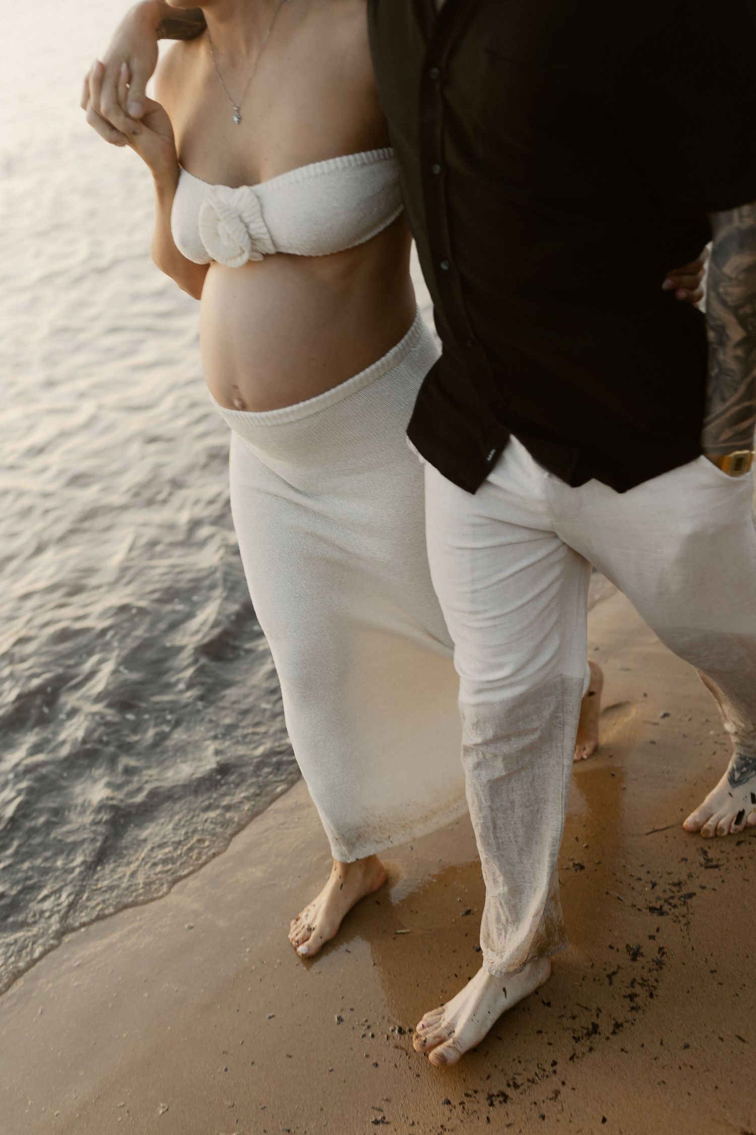 A couple in white beachwear stands at the water's edge during golden hour, creating an intimate coastal moment.