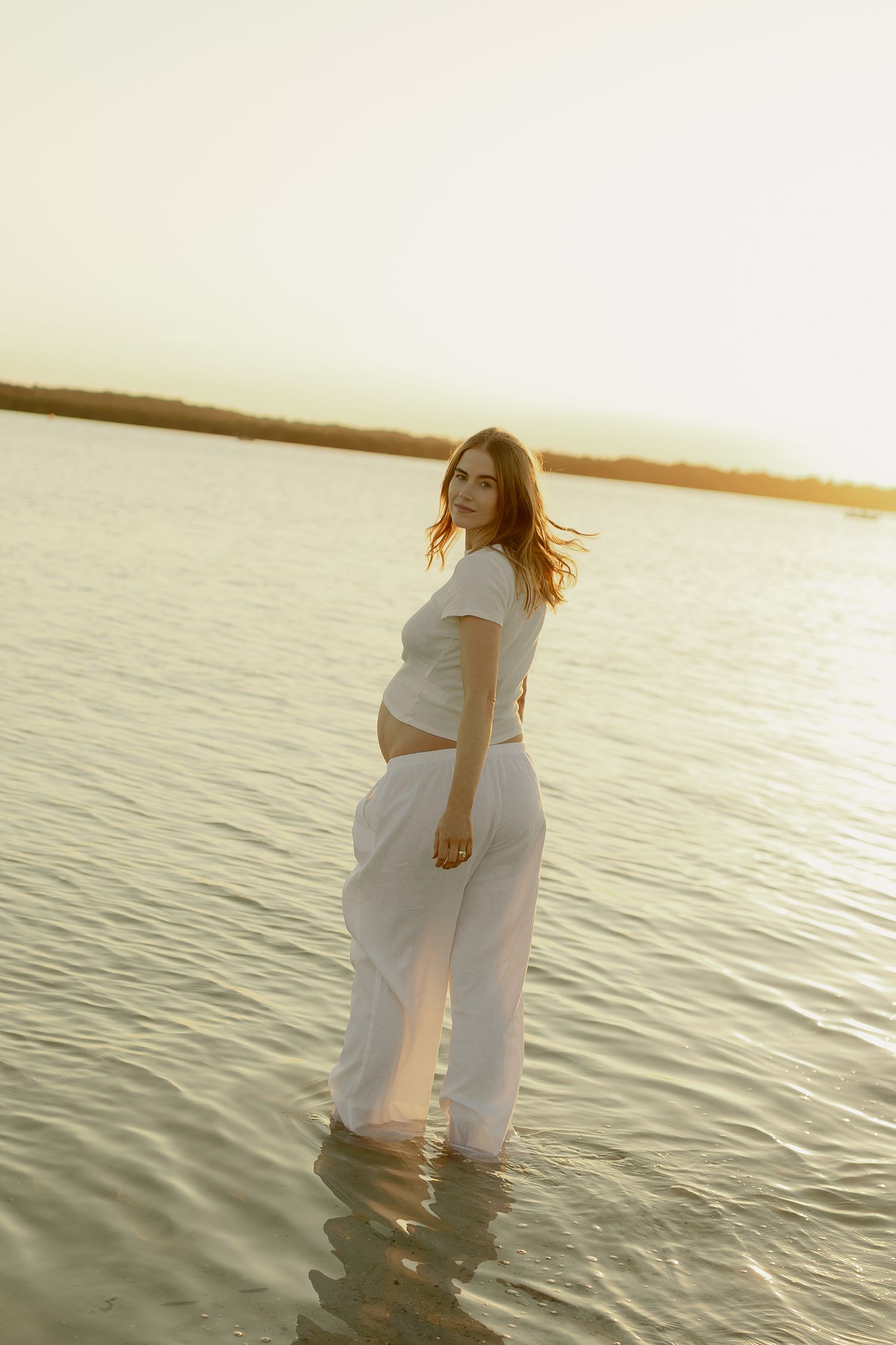 Pregnant woman in white crop top and pants standing in shallow water at golden hour sunset.