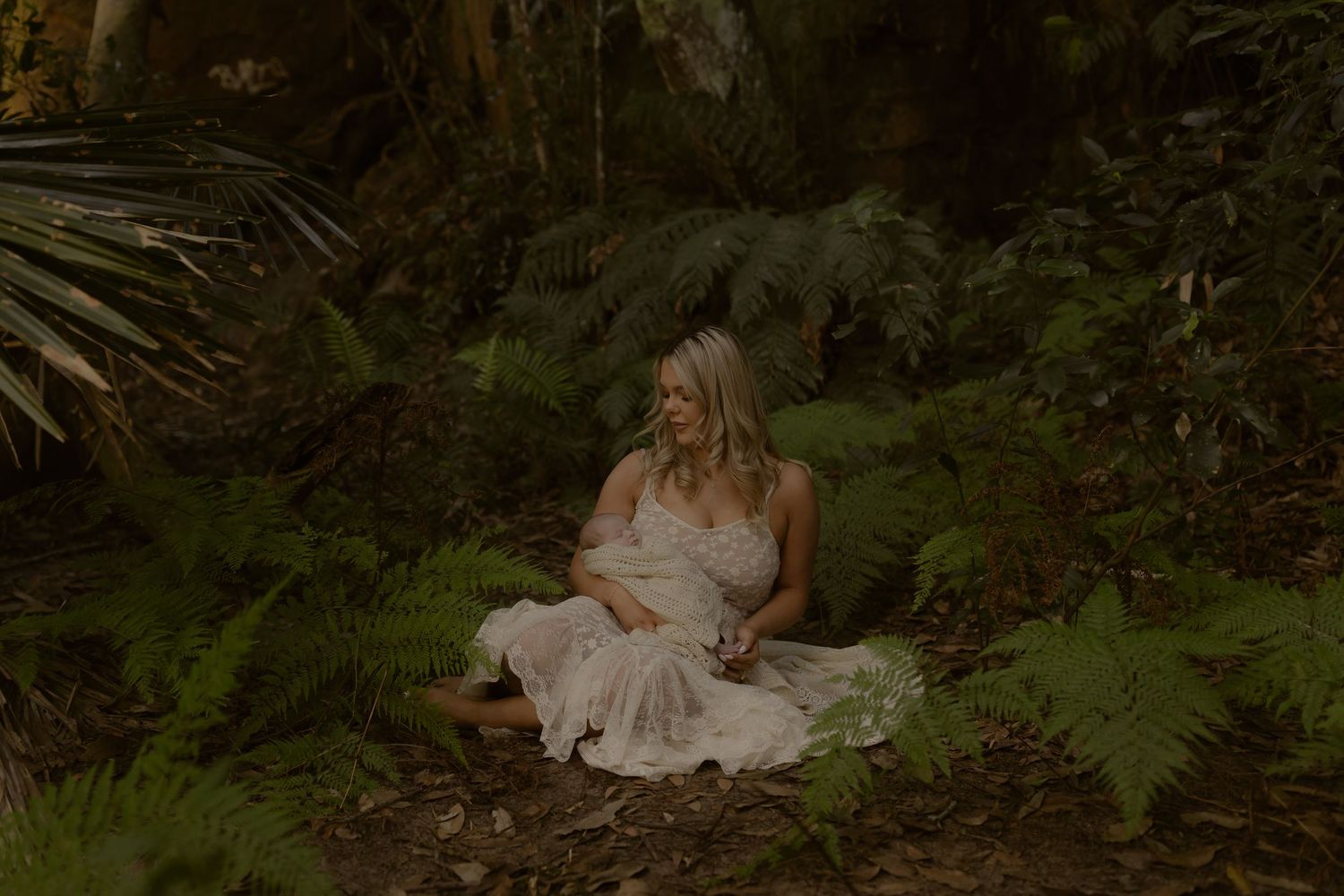 Pregnant woman in white lace dress sitting peacefully among lush green ferns in a serene forest setting.