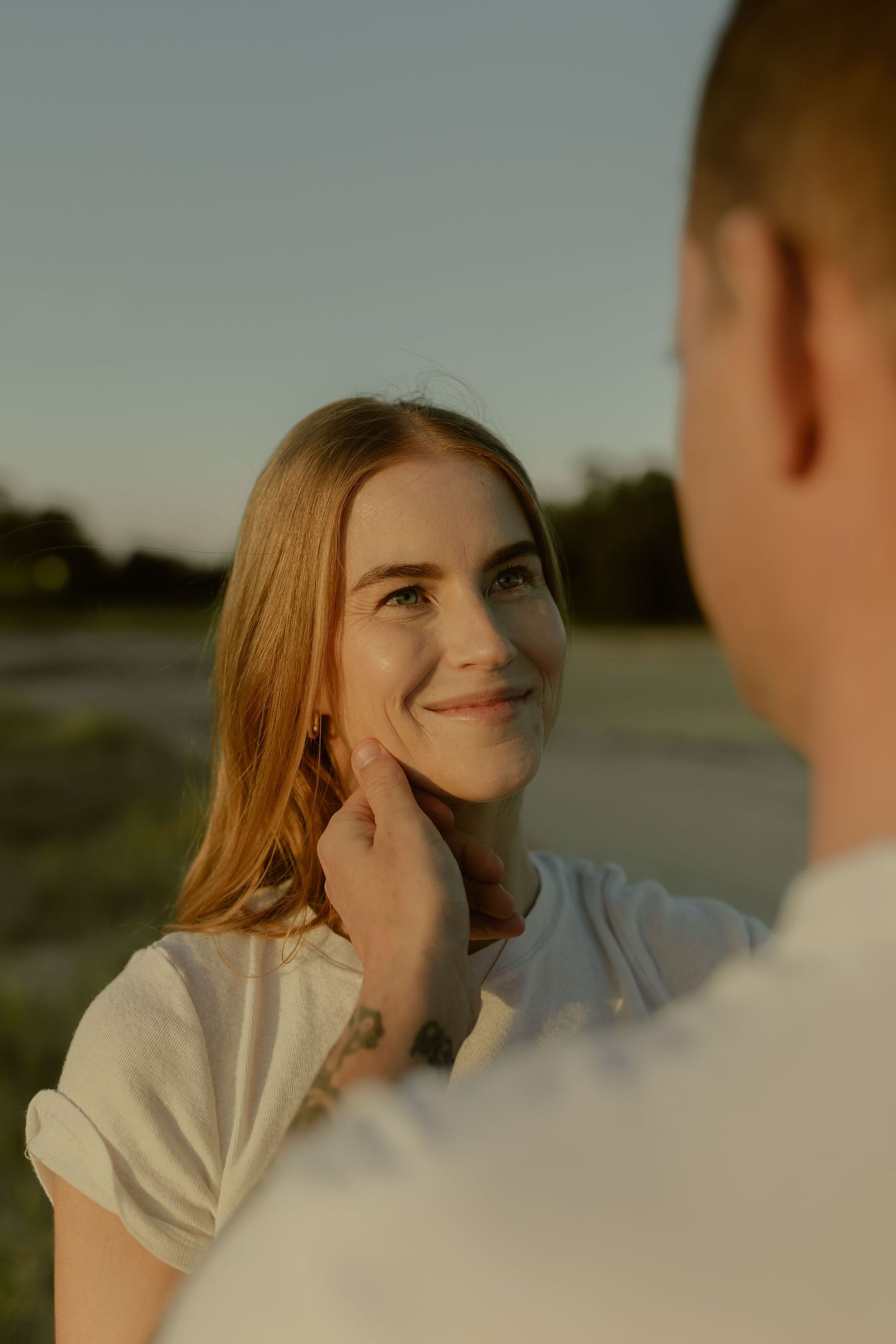 Young woman with long auburn hair smiling warmly as man gently touches her face during golden hour outdoors.