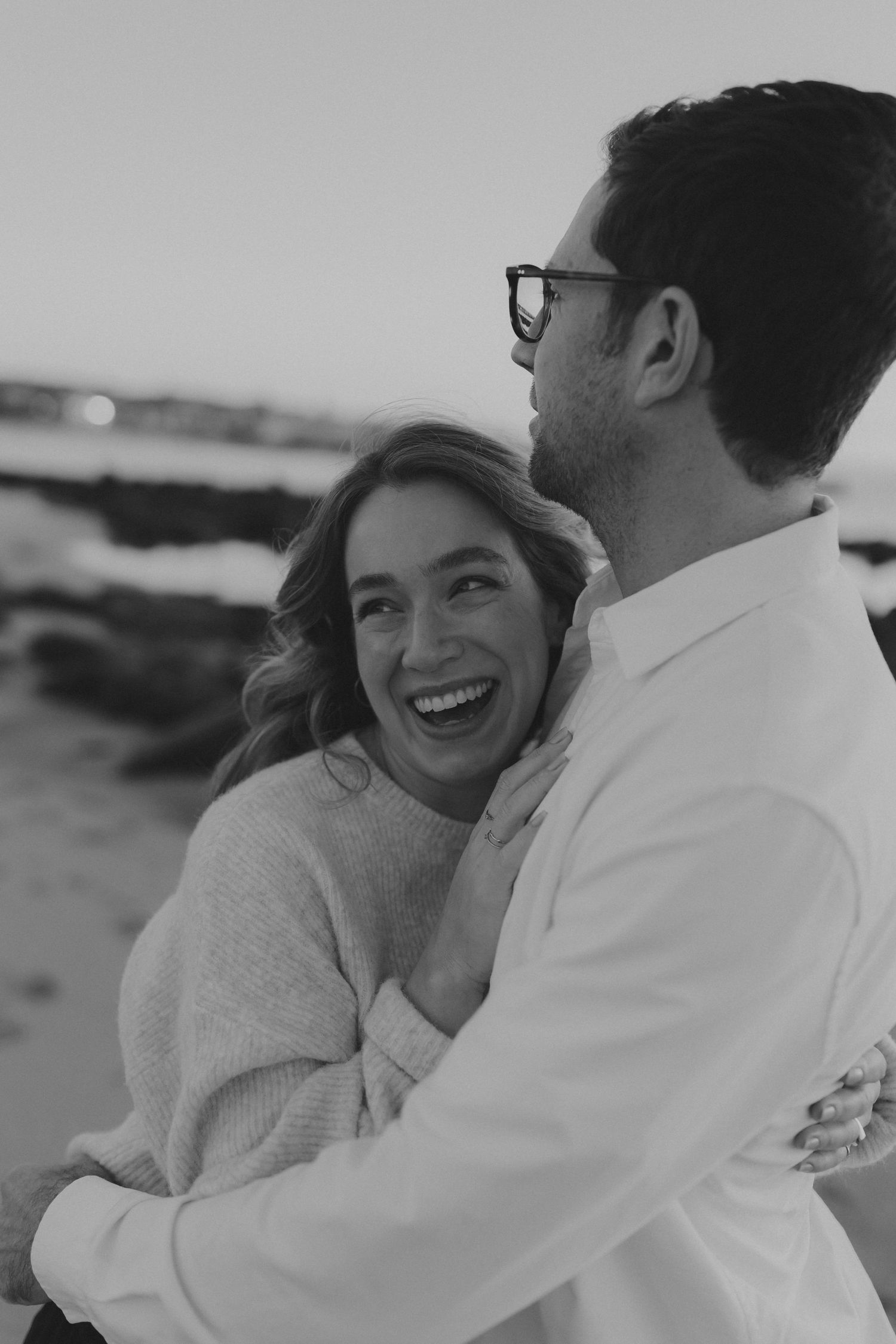 Black and white photo of joyful couple embracing on beach, woman laughing while holding man in glasses.