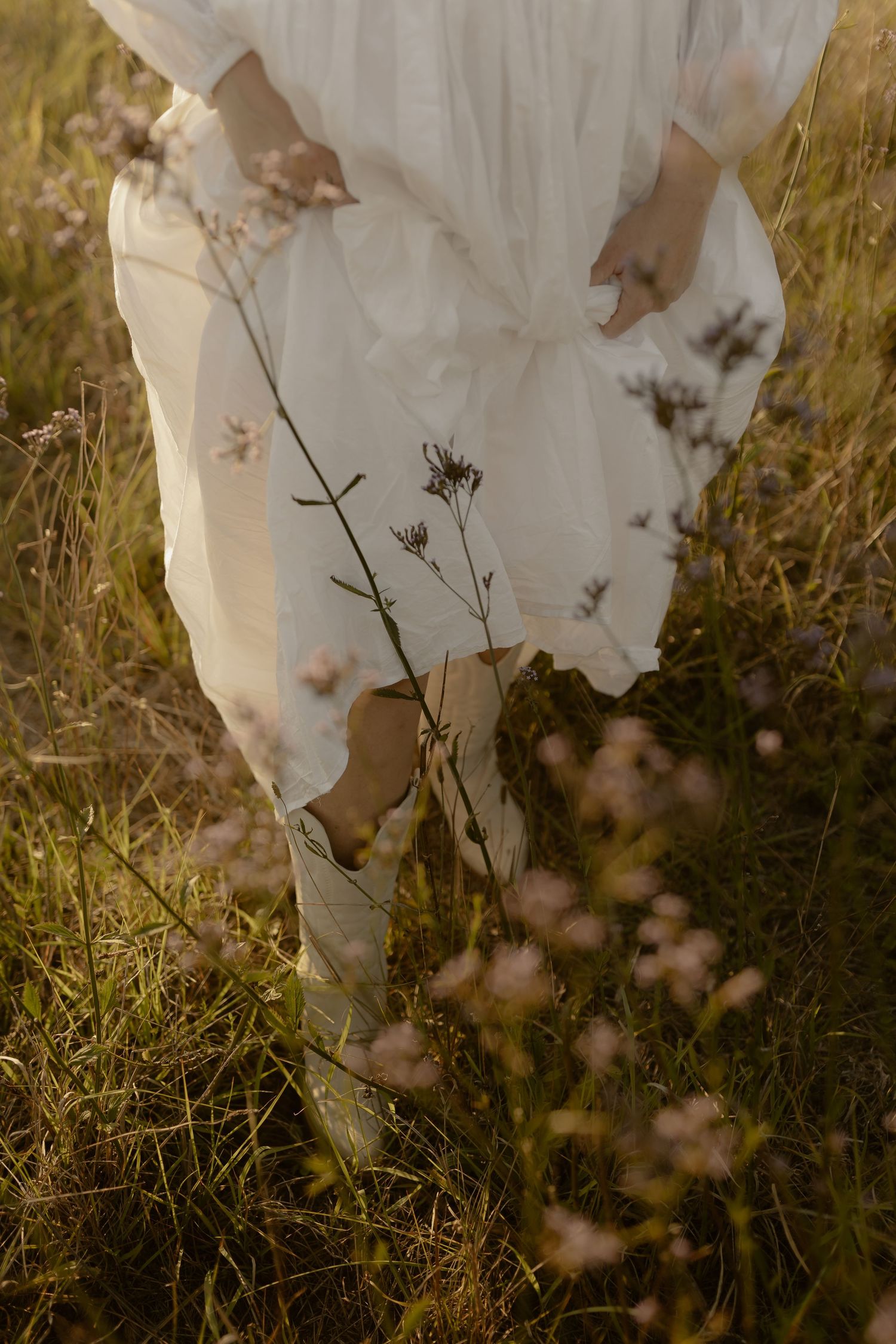Close-up view of a white flowy dress against tall grass and wildflowers in golden sunlight.