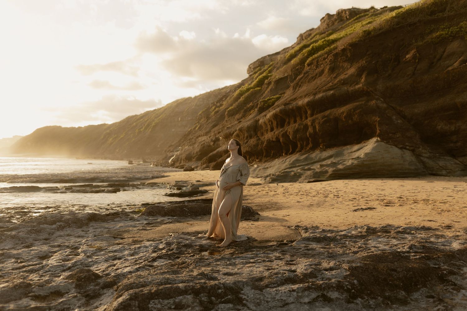 A figure in flowing fabric stands on a rocky beach at sunset with dramatic coastal cliffs in the background.