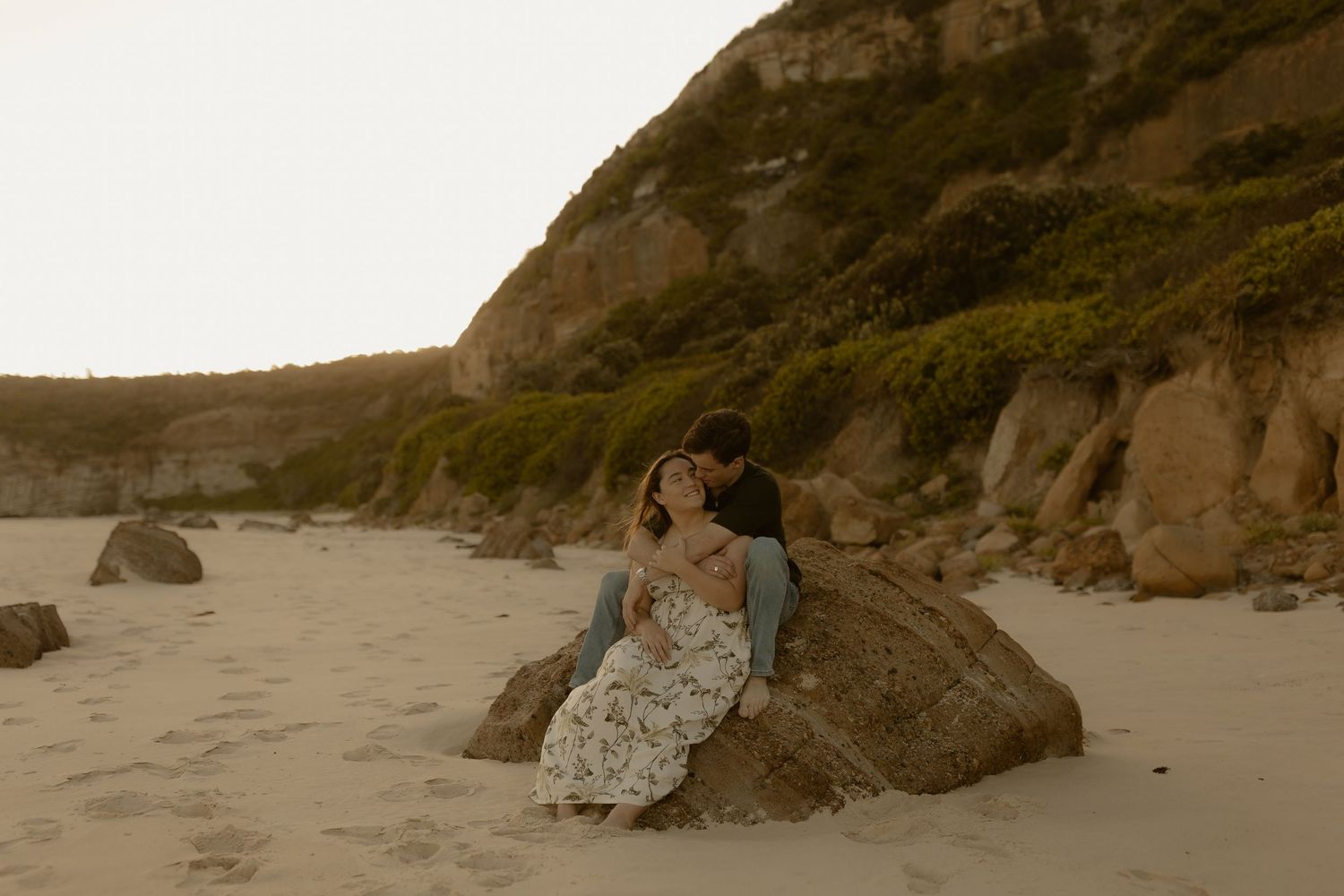 A couple sits together on a large rock at sunset on a secluded beach with cliffs in the background.
