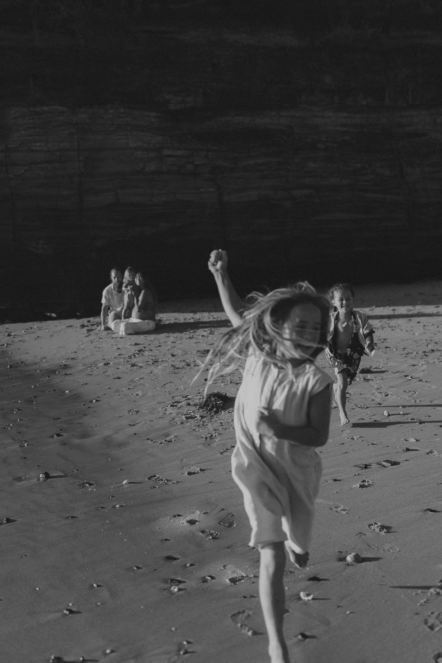 A black and white photo of a person dancing joyfully on a beach with others in the background.