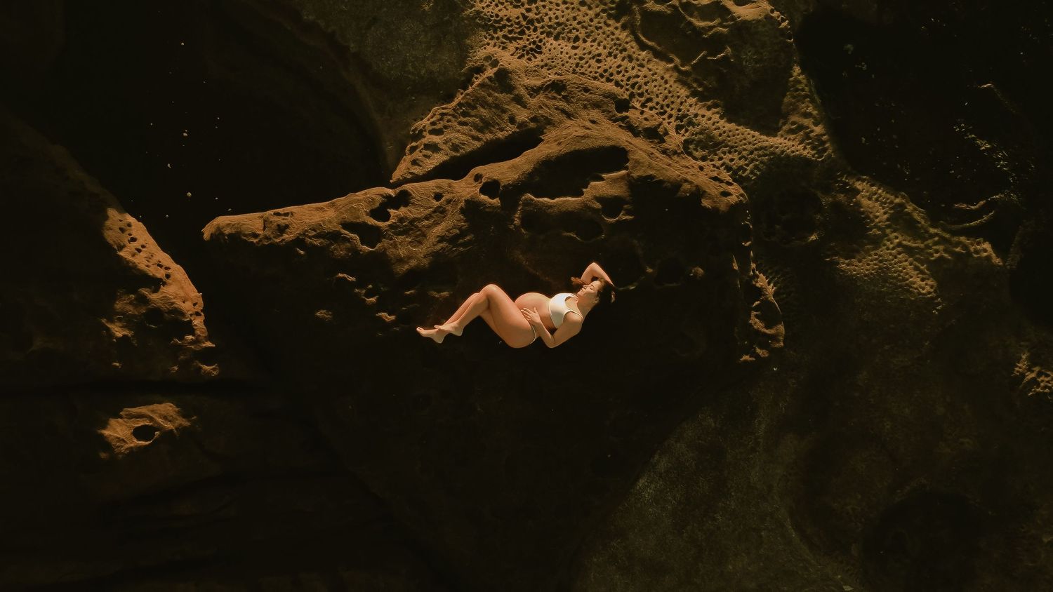 A person floats in dark water surrounded by dramatic rocky formations and dramatic shadows in a natural cave setting.