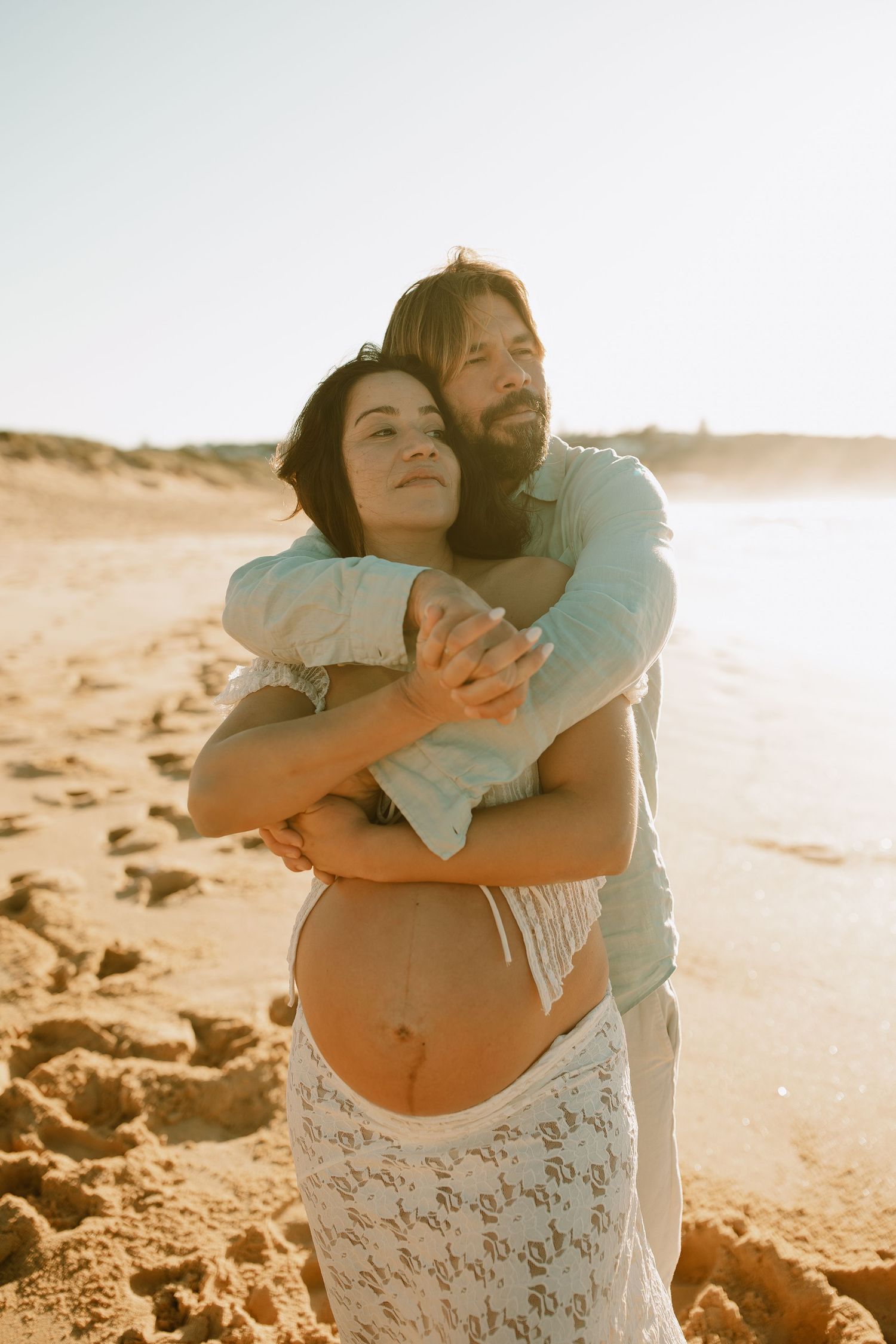 Couple shares tender moment during beach maternity photoshoot at sunset.