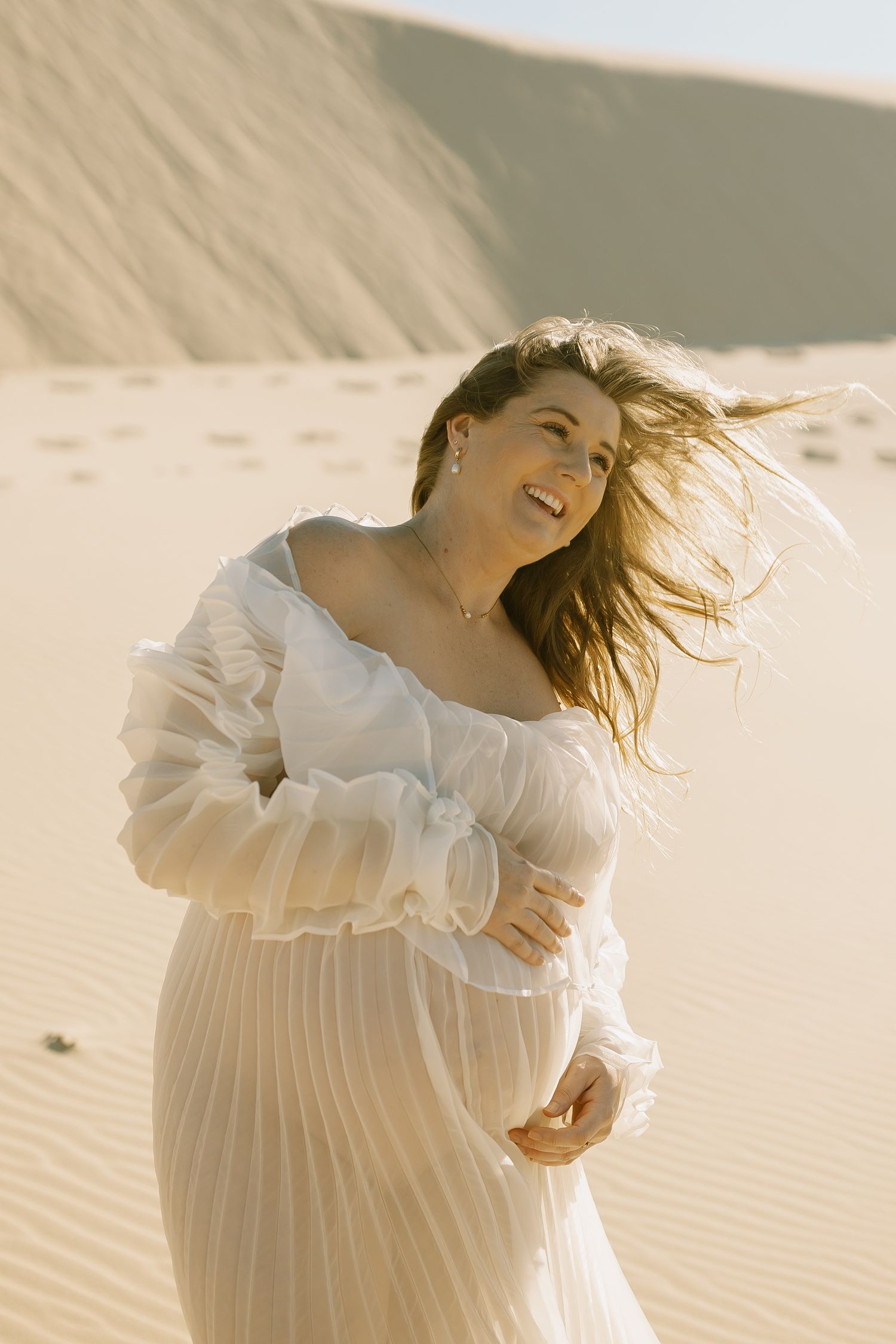 A person wearing white ruffled clothing poses dramatically against sandy desert dunes at sunset.