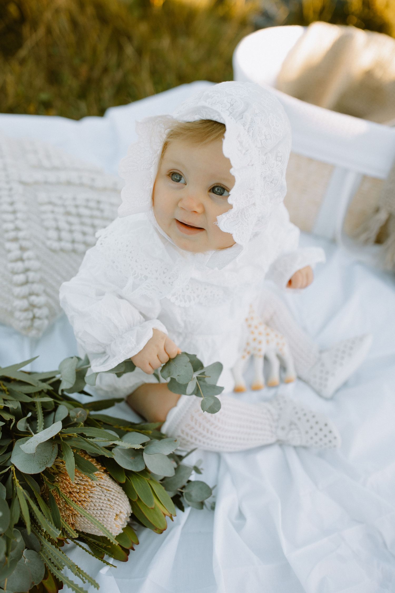 A baby wearing a white knit bonnet sits with greenery and floral arrangements on a white textured blanket outdoors.