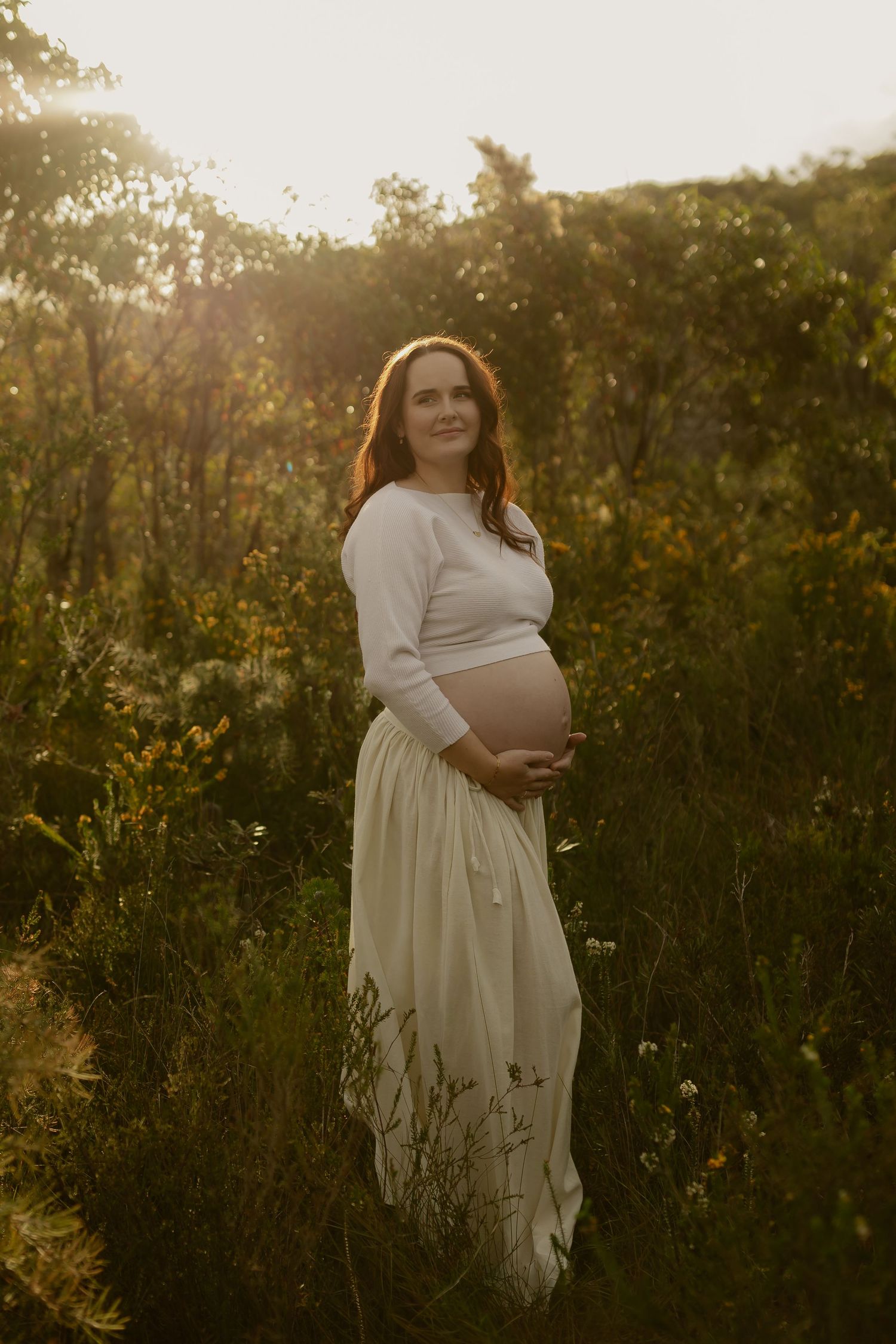 Pregnant woman in white outfit cradling baby bump in golden hour sunlit field.