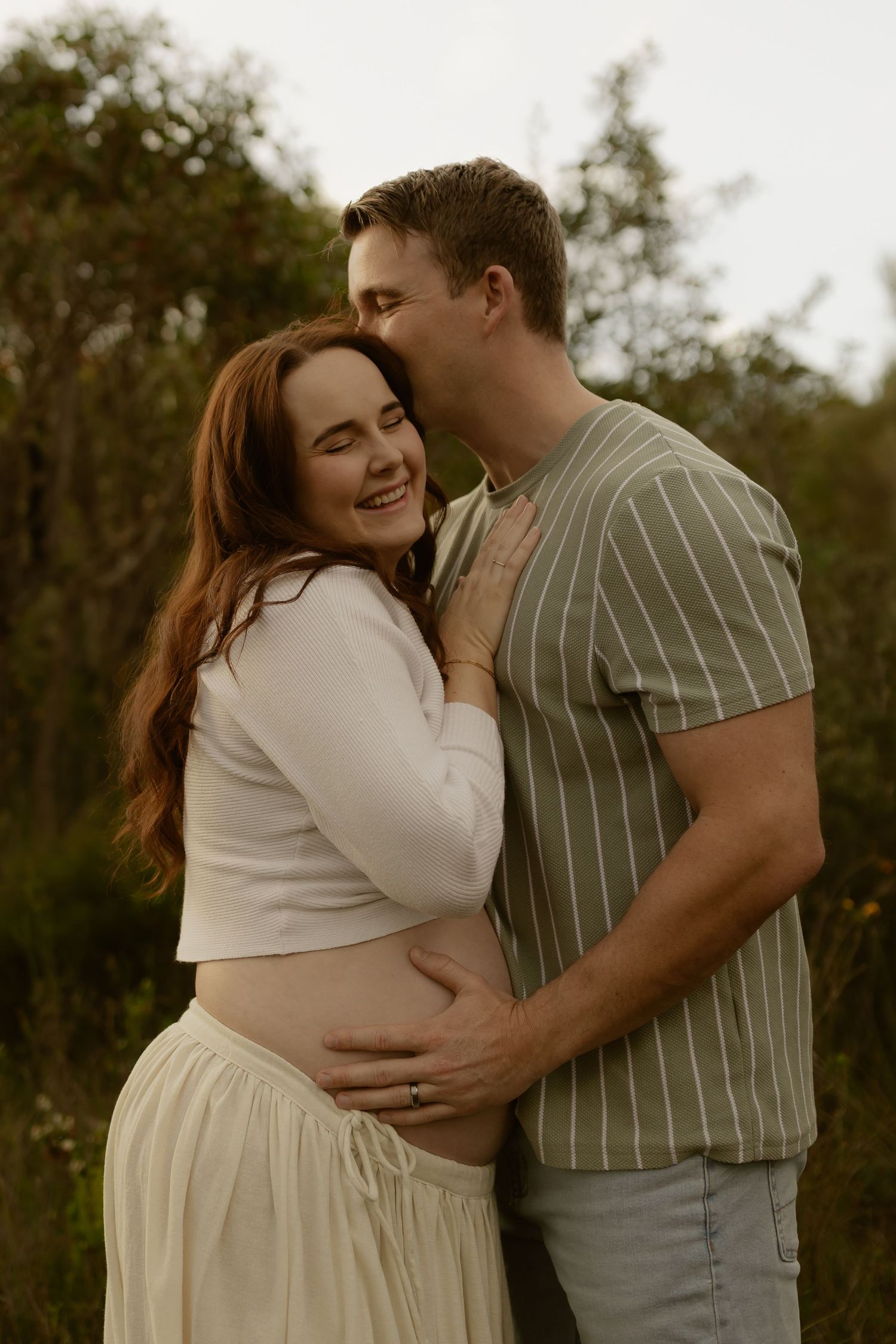 A couple embraces outdoors, man kissing woman's temple as she smiles, her red hair flowing in a maternity photo session.