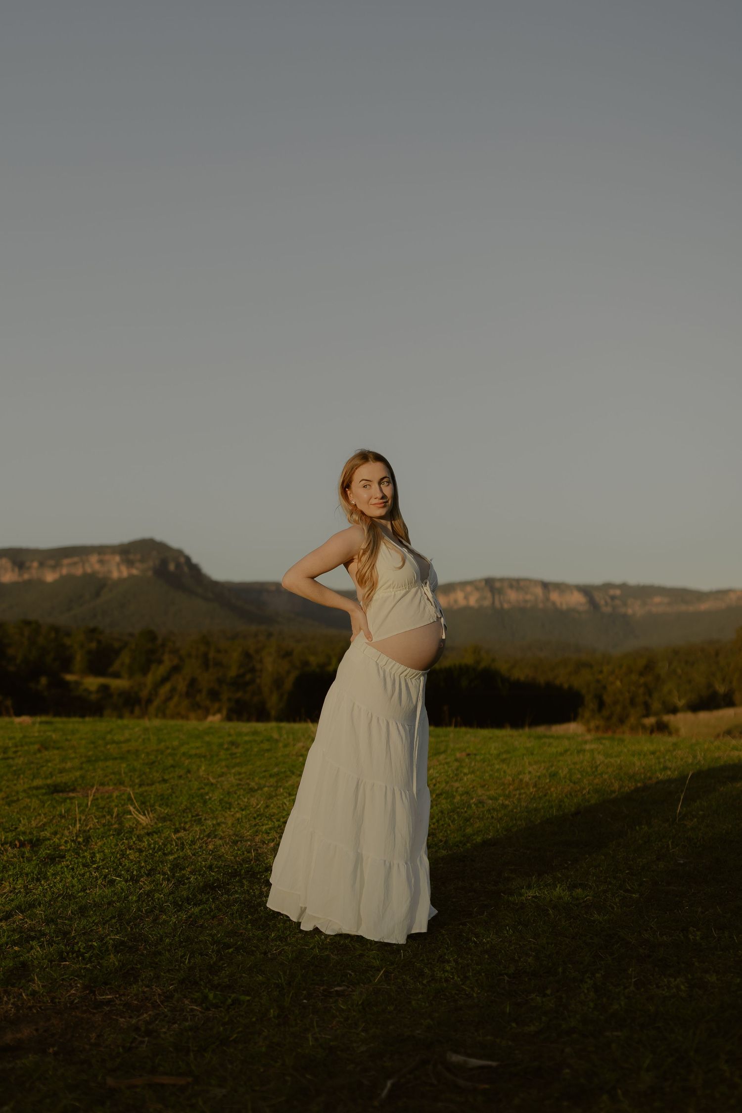 Pregnant woman in white maxi dress standing on green grass with mountain backdrop at golden hour.