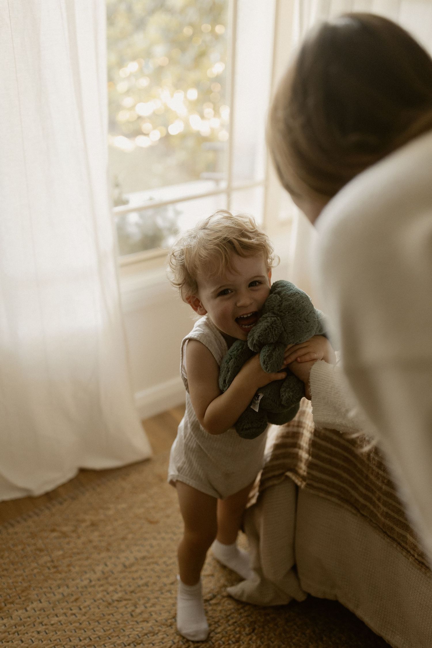 A toddler in white clothes hugging a stuffed toy by a curtained window.