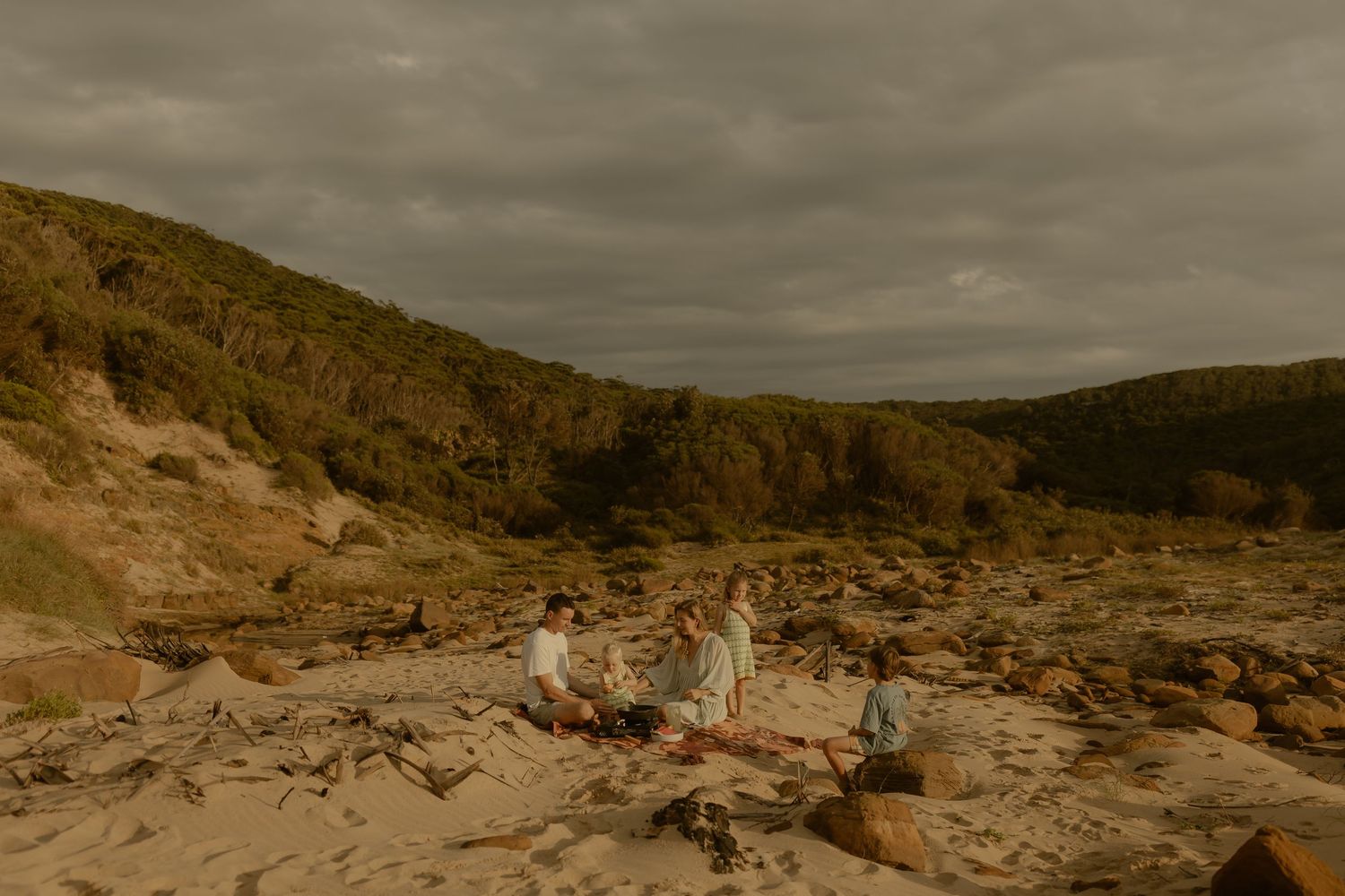 Two small figures rest on rocky coastal terrain under dramatic cloudy skies.