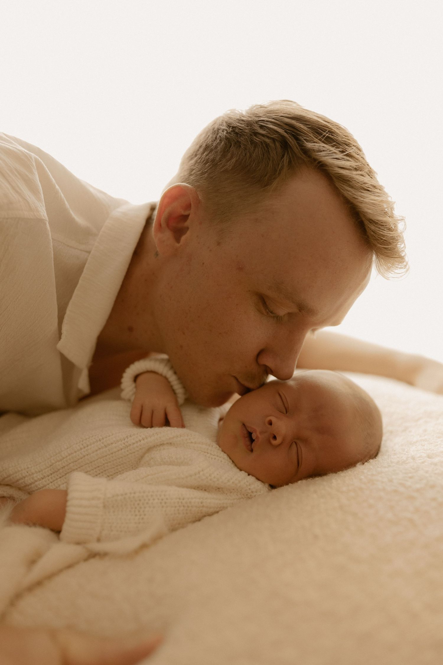 Tender sepia-toned moment showing gentle affection between parent and infant on white bedding.