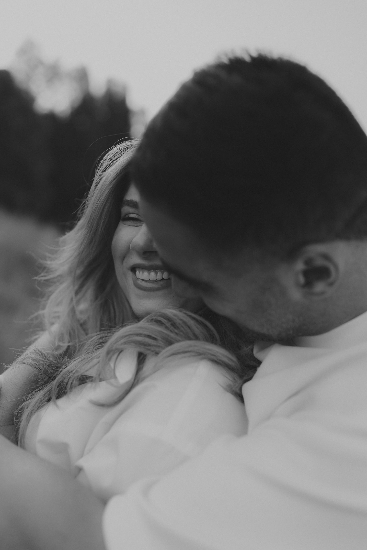 Black and white photo of laughing pregnant woman being embraced by partner in outdoor field setting.