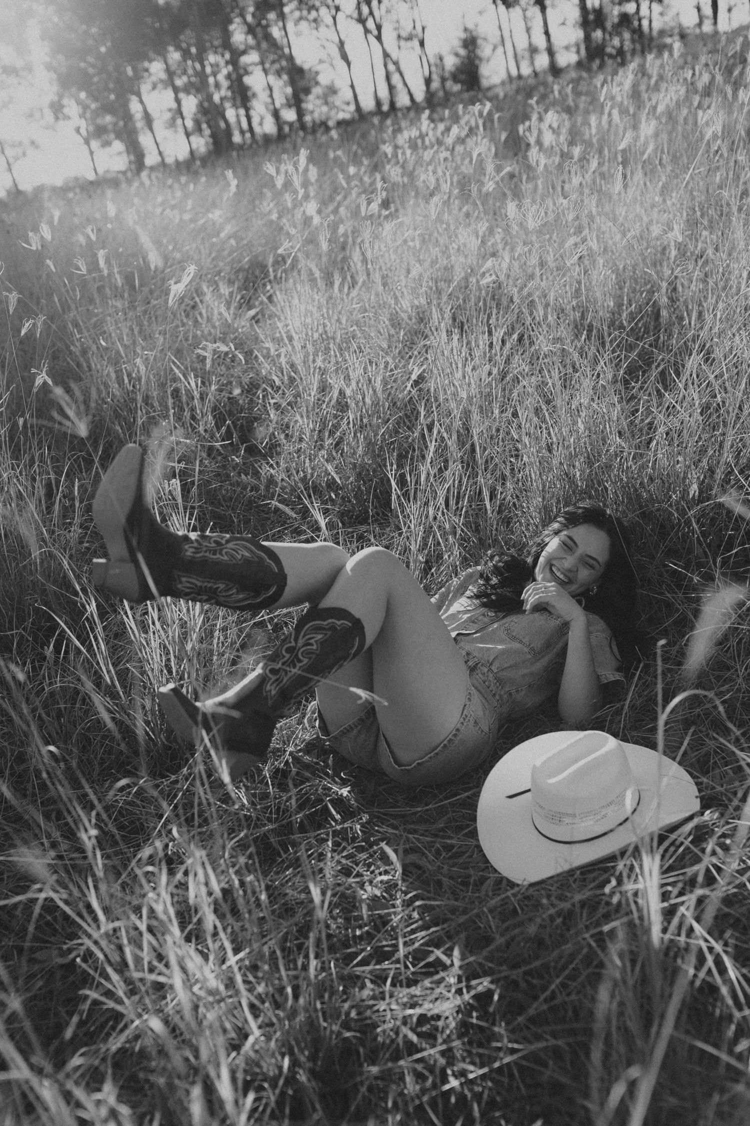 Black and white photo of woman lying in tall grass field wearing cowboy boots with hat beside her.