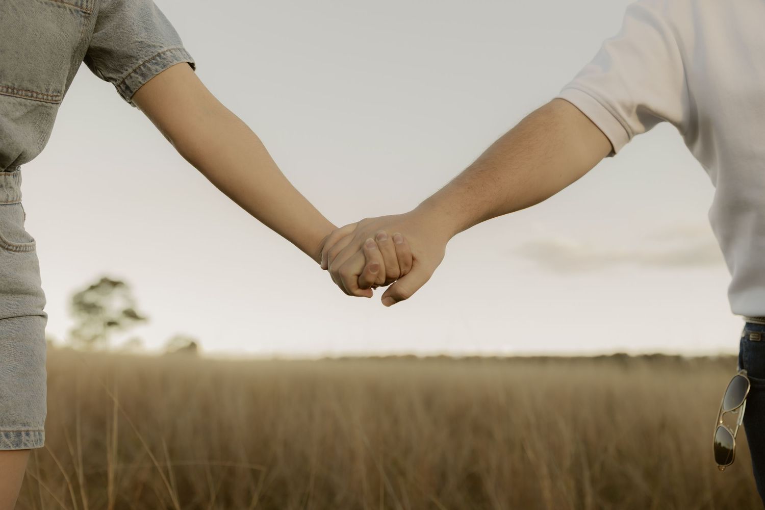 Close-up of couple holding hands together against golden grass field and soft sunset sky.