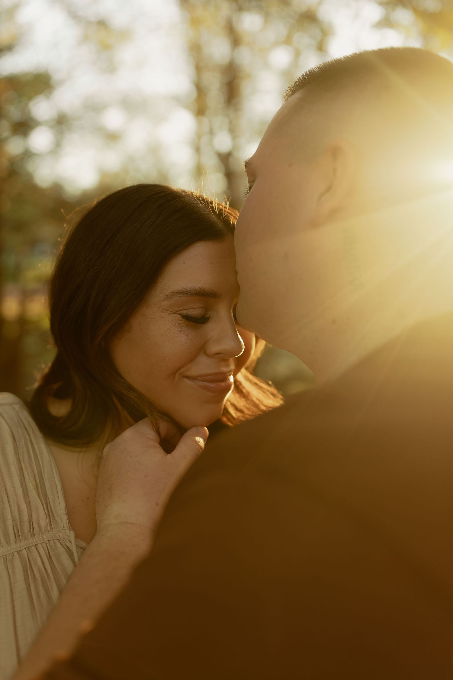 A couple shares a tender moment at golden hour, with warm sunlight glowing behind them as a man kisses a smiling woman.