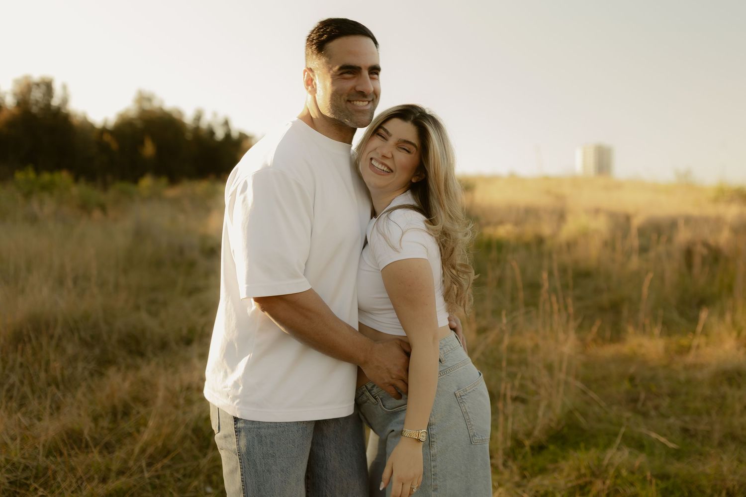Happy couple in white t-shirts embracing in a golden field at sunset, smiling warmly together.
