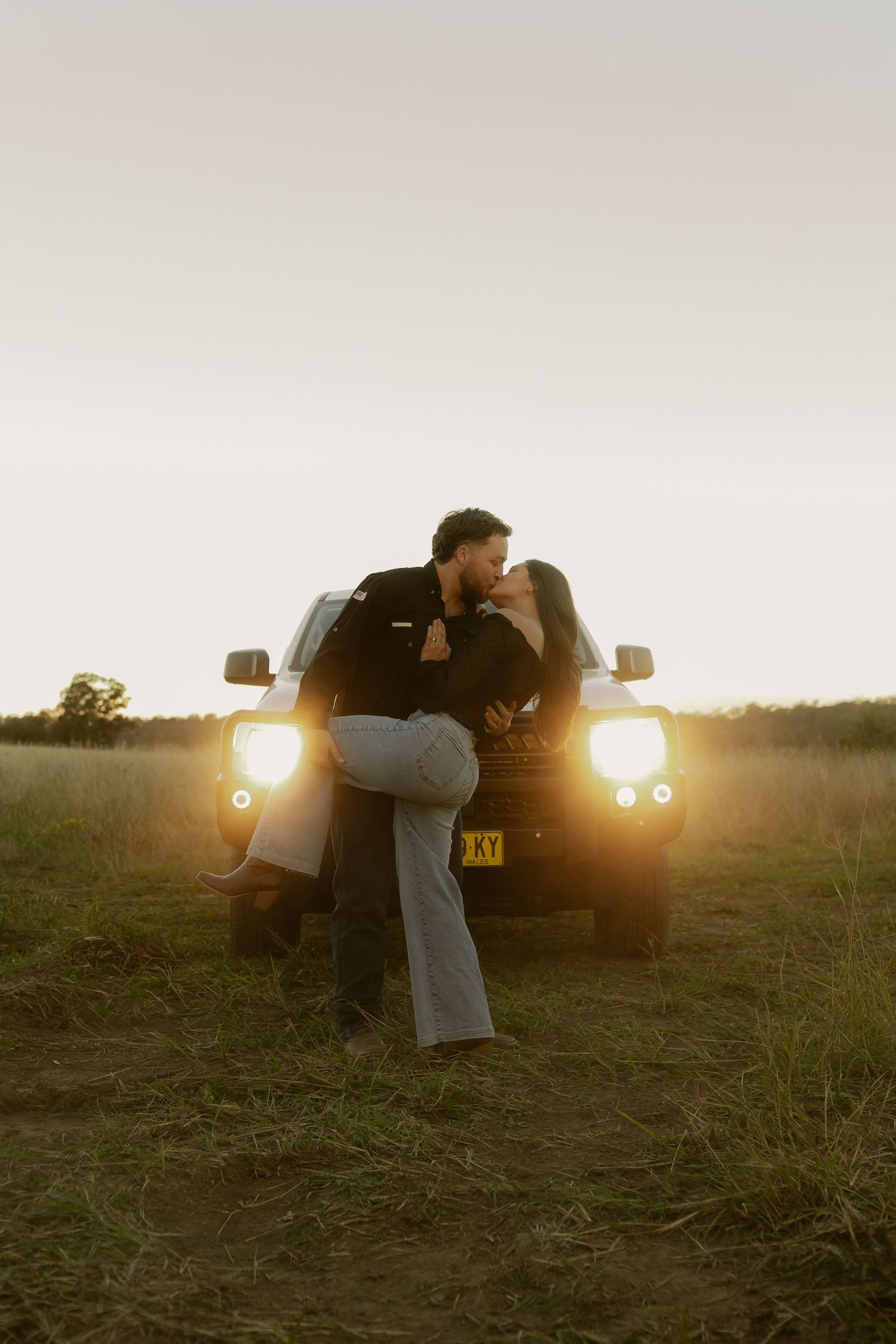 Romantic couple sharing a kiss in front of glowing car headlights at dusk in an open field.